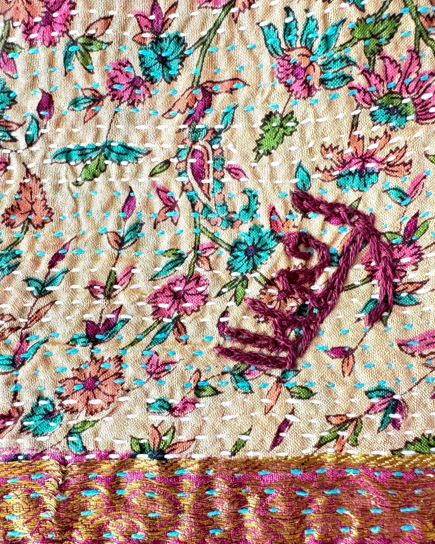 Colorful floral embroidery on beige fabric, featuring pink, blue, and green flowers. A shiny pink and gold patterned border adds a vibrant touch.