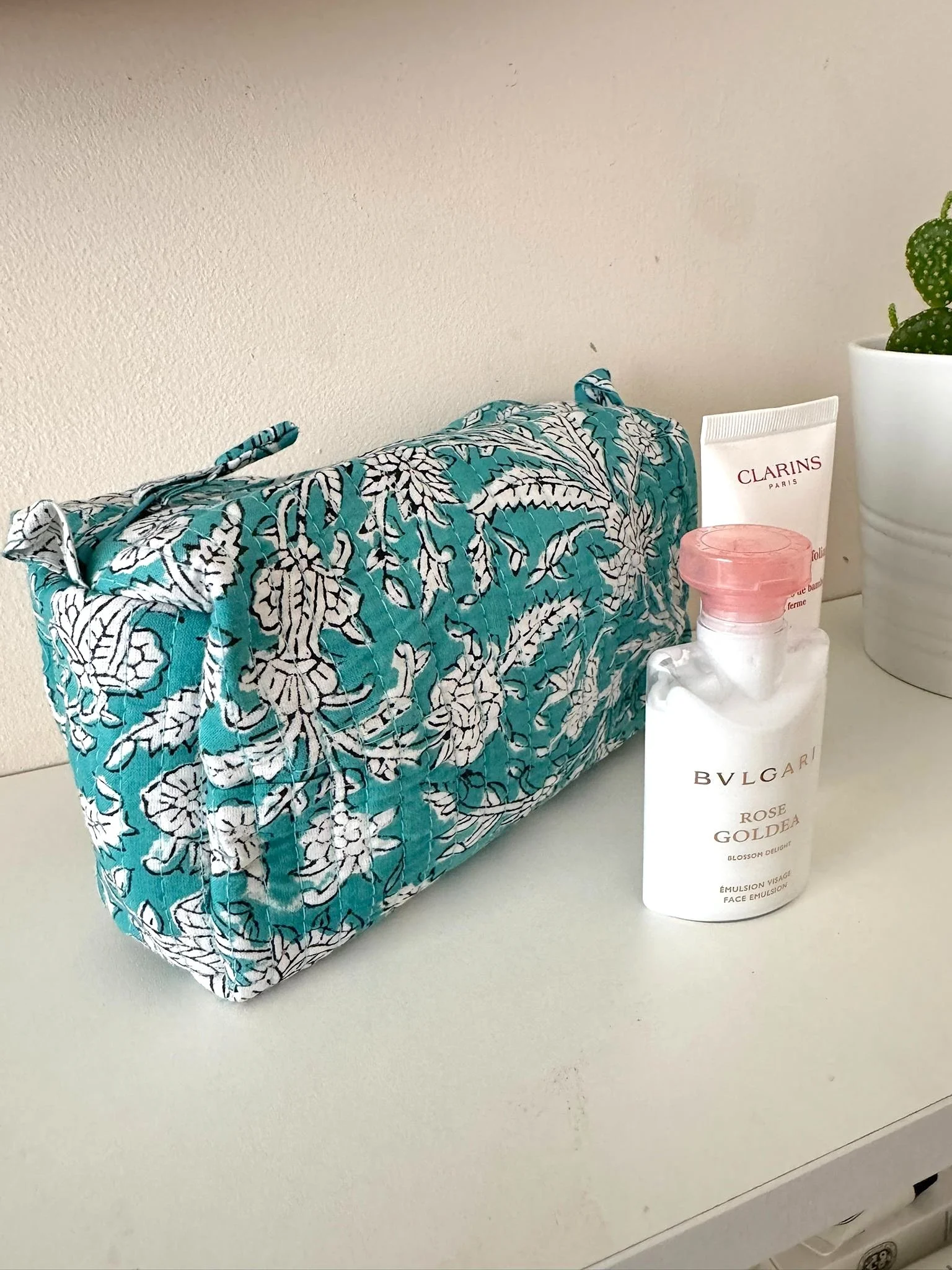 Colourful wash bag in turquoise and white resting on a bedroom shelf with luxury travel sized toiletries in front