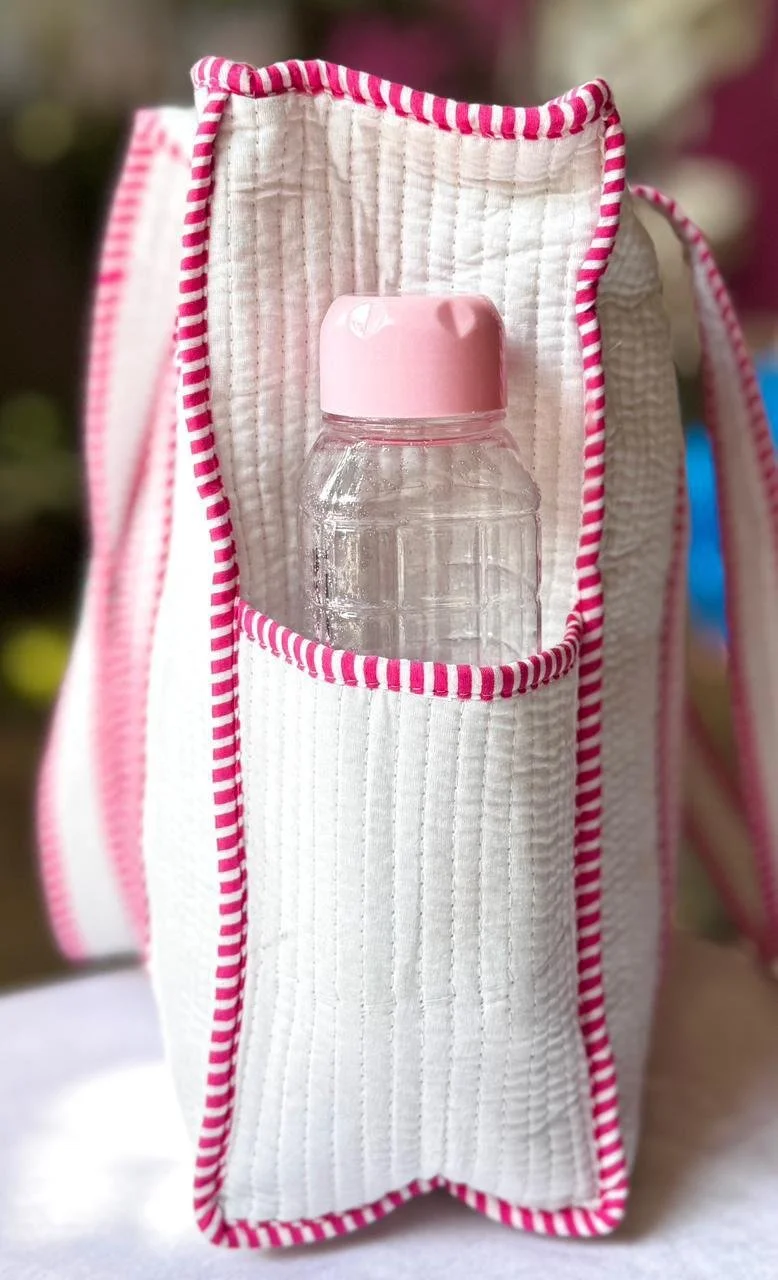 Close up of a bottle with a pink lid placed inside the external pocket of a white and pink striped handmade quilted cotton tote bag.
