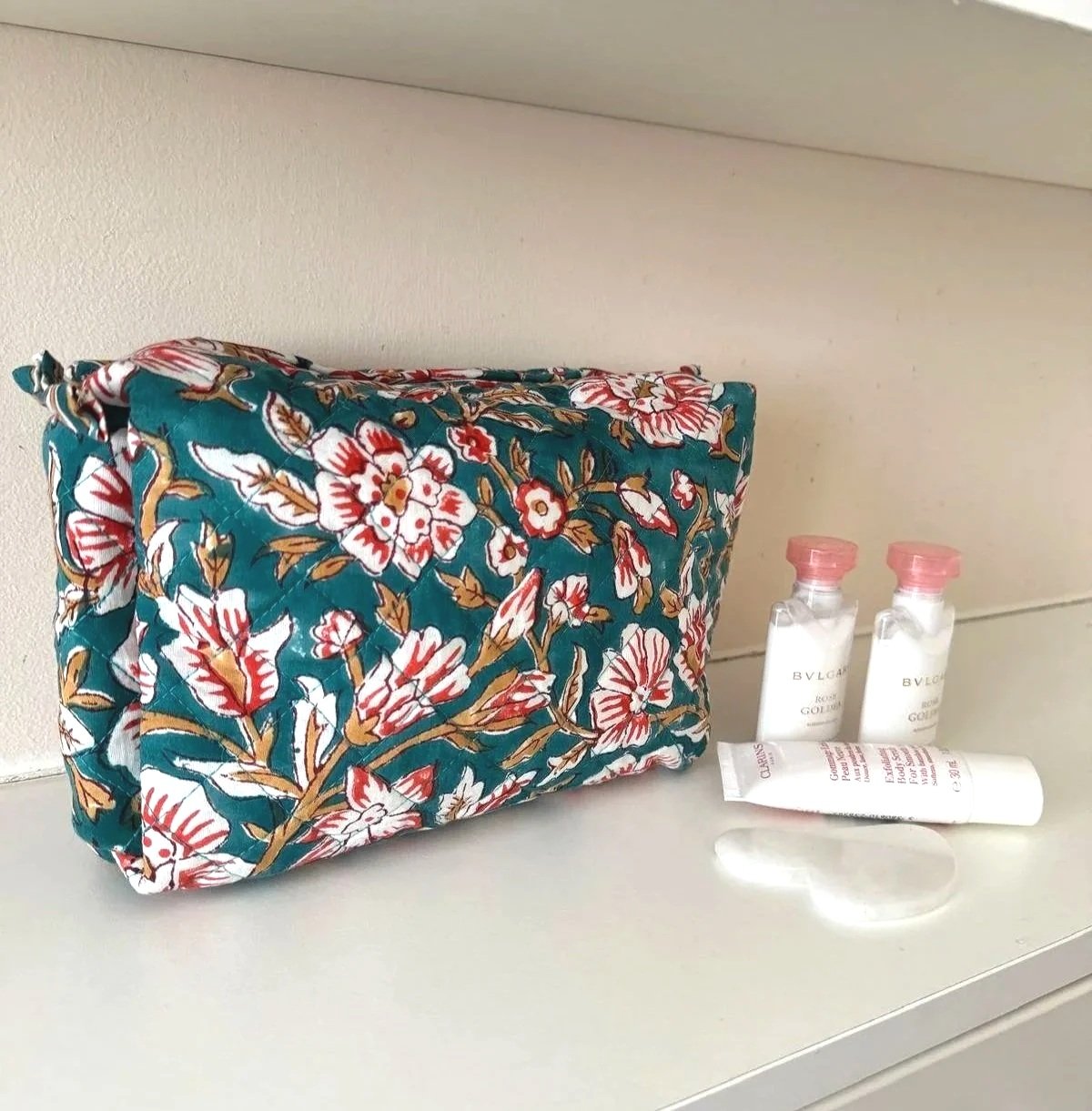 Large travel wash bag resting on a white shelf with travel sized toiletries in front, the wash bag is a teal colour with large white and red flowers and is hand block printed