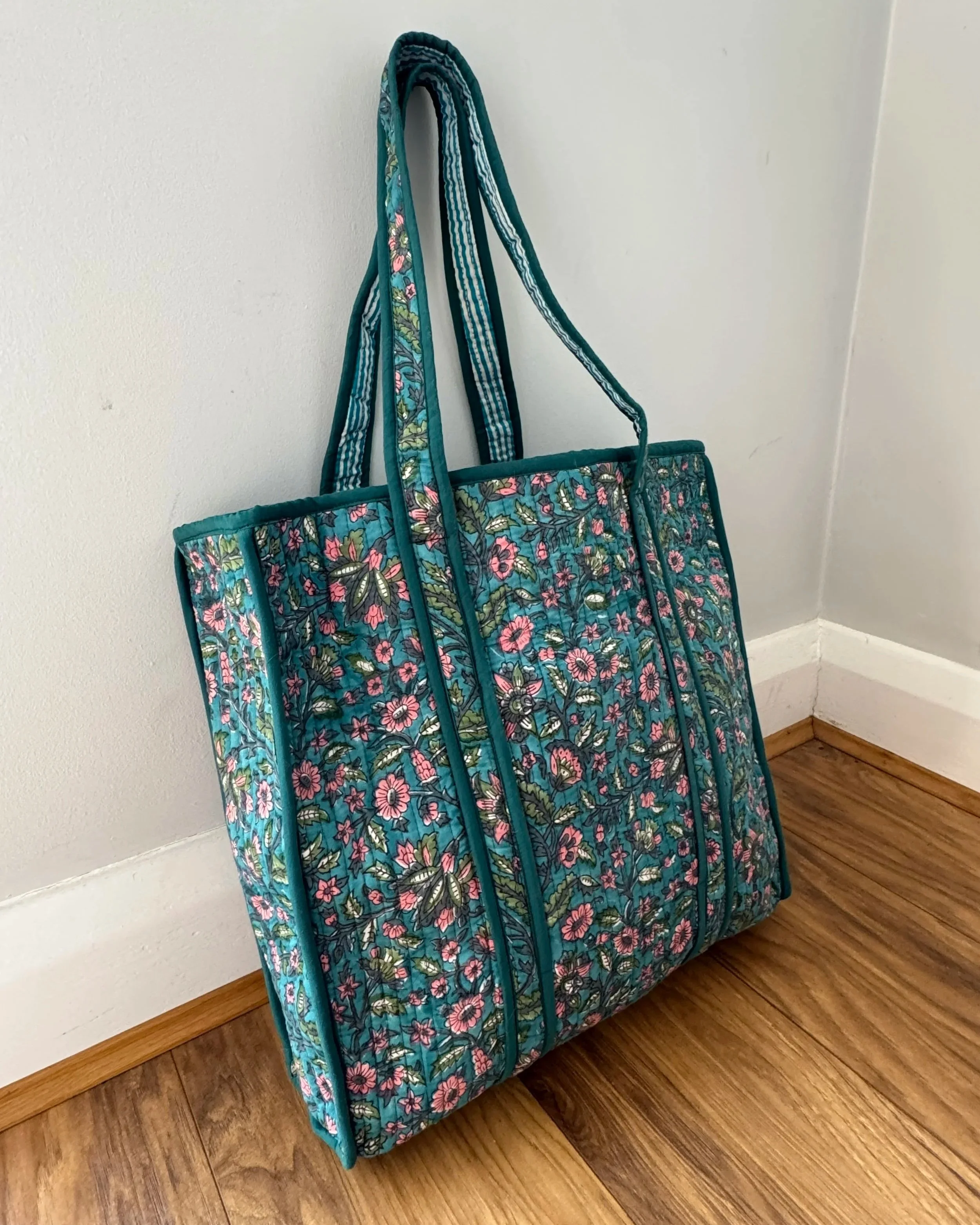 Indian hand block printed floral quilted cotton bag with pink and green flowers and contrasting striped shoulder straps, leaning against a light grey wall and wooden floor