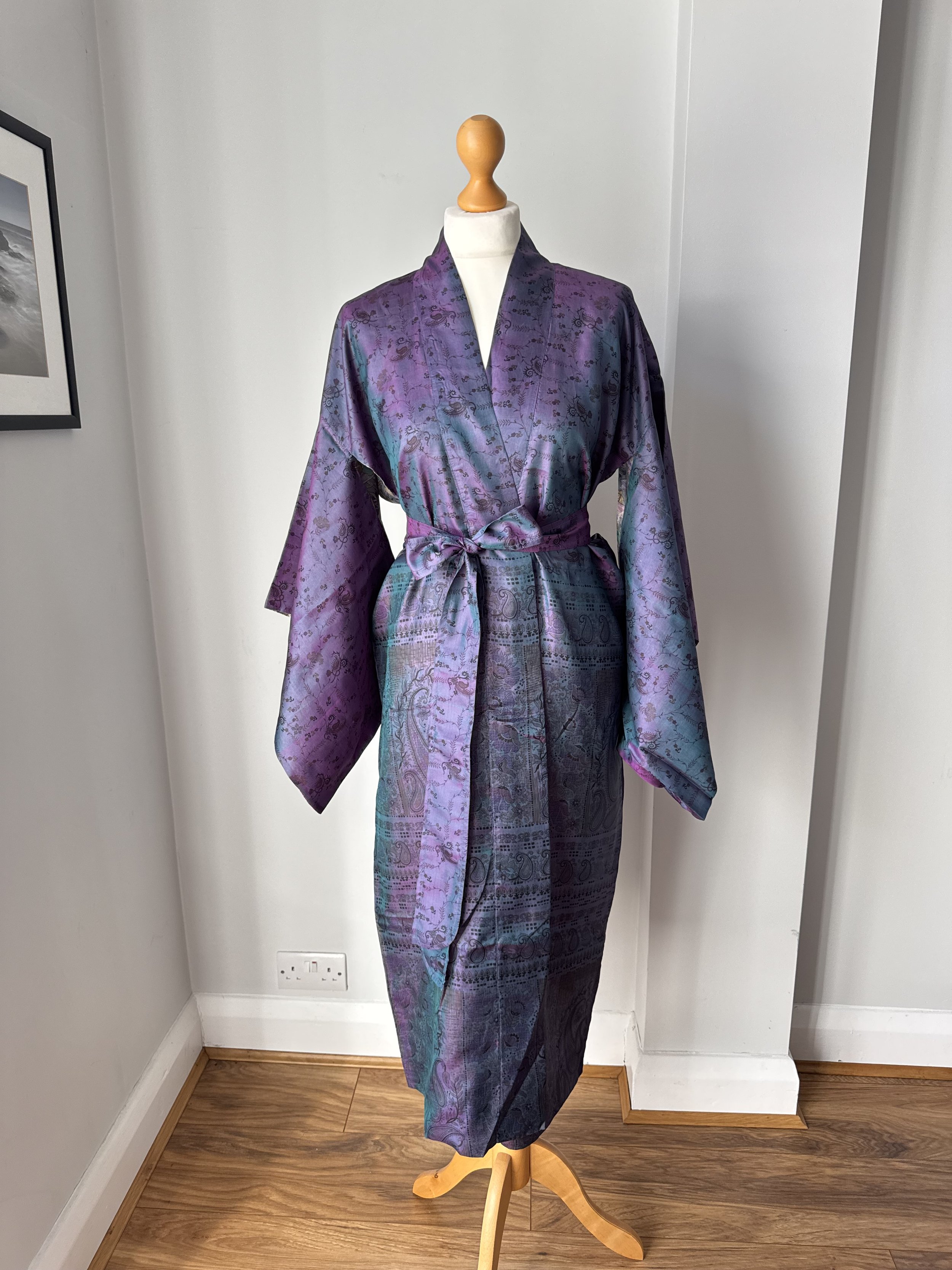 Long kimono robe in two-tone purple and blue paisley and floral print, fastened at the waist with a matching tie belt, worn on a dressmaker's mannequin against a grey wall