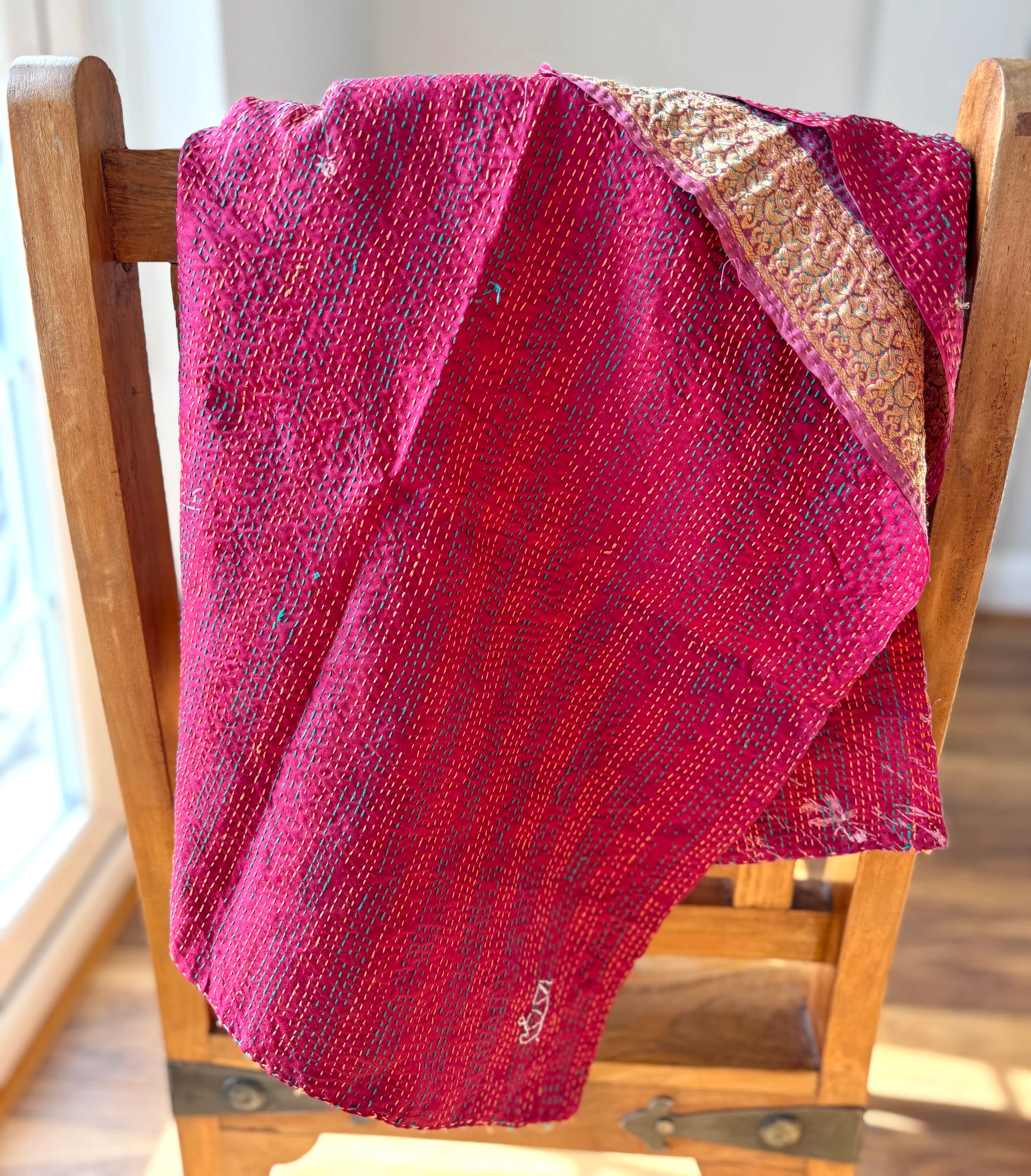 A vibrant, patterned magenta silk scarf quilted with intricate embroidery hangs over a wooden chair in a sunlit room