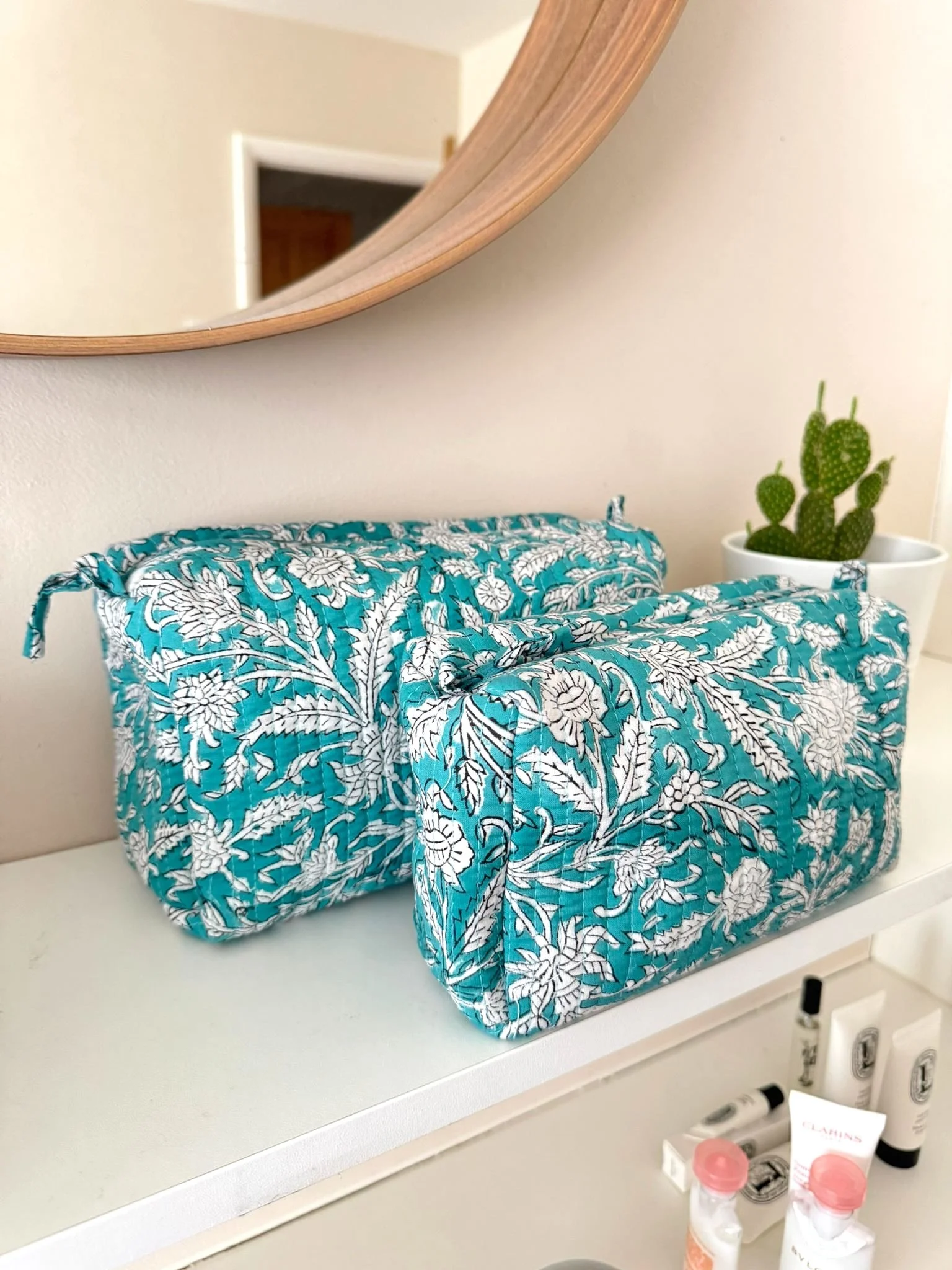 Two colourful turquoise and white cosmetic bags resting on a white shelf, the wash bags are hand block printed in a floral print