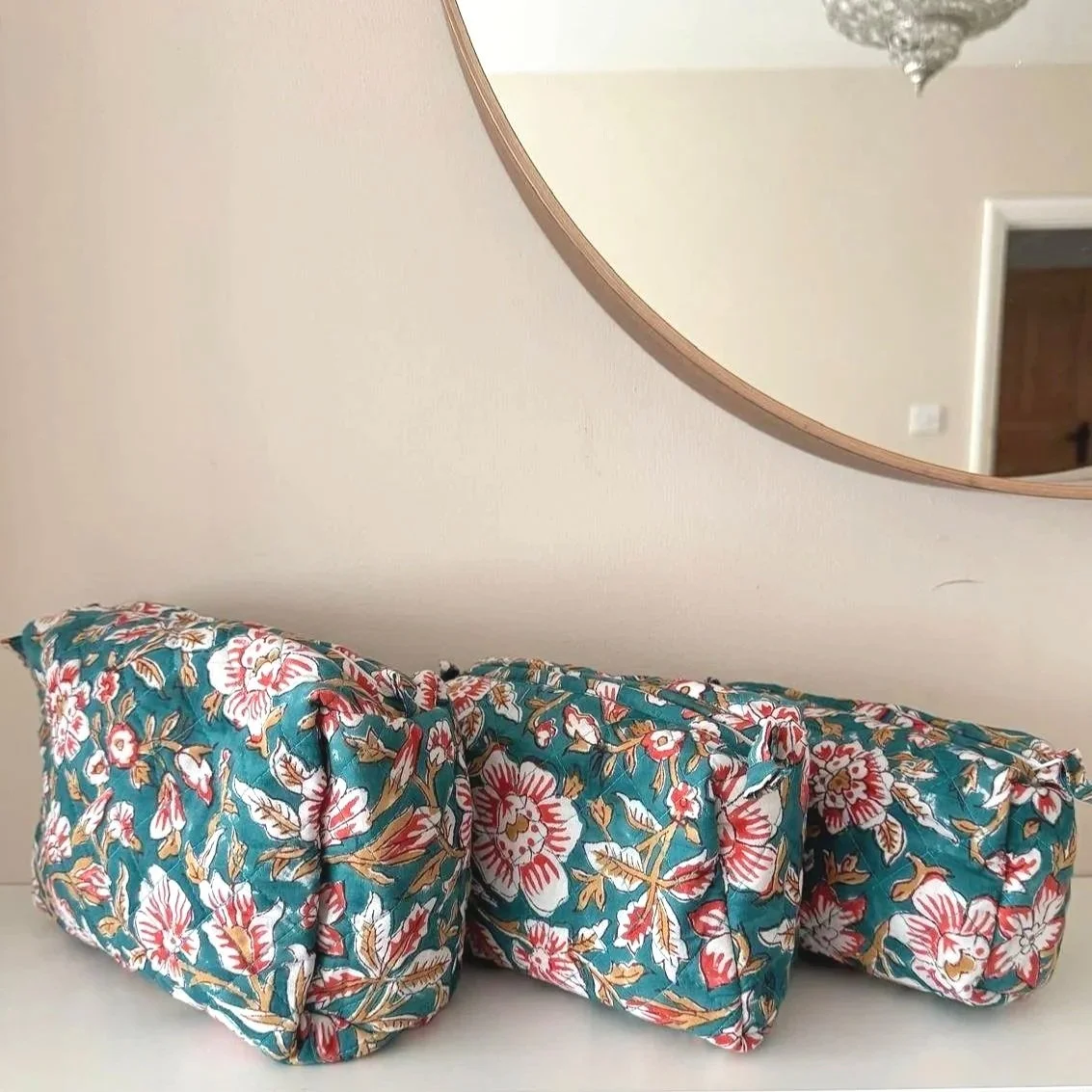 Set of 3 hand block printed teal wash bags resting on a white shelf. The wash bags are teal with large white flowers and red highlights