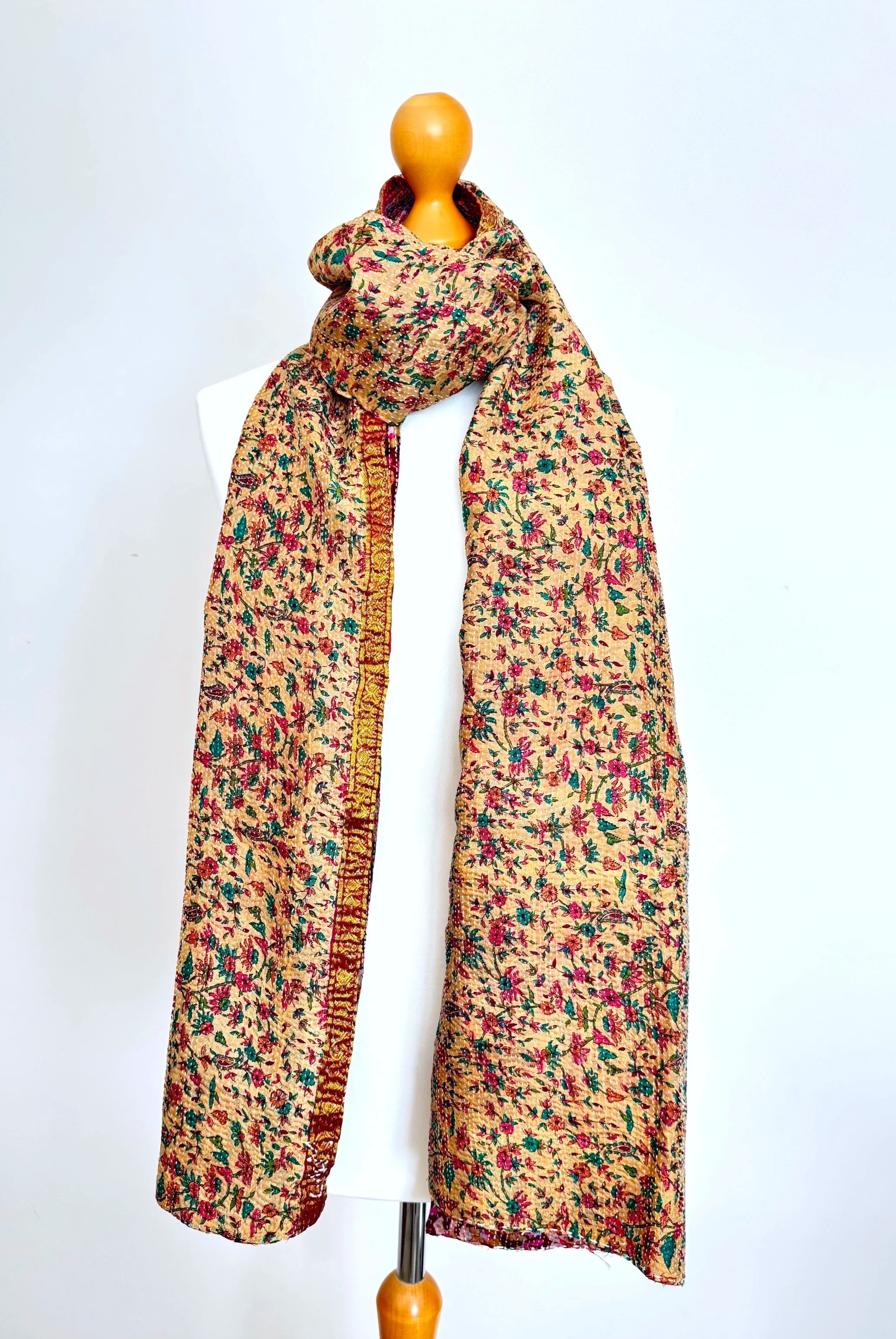 Intricate floral embroidery with vivid pink, teal, and green threads on a tan fabric. A gold and magenta woven border runs along the top edge.