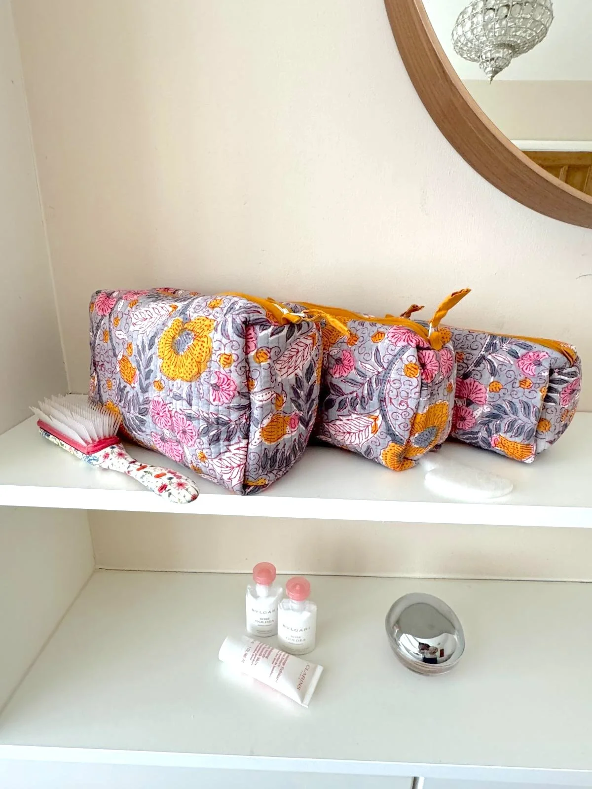 Set of 3 colourful  quilted cotton wash bags with a floral print in shades of pink, yellow and grey, resting on a white shelf surrounded by toiletries