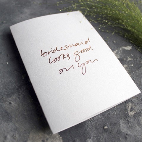 A white bridesmaid card on an angle against a grey backdrop that reads, bridesmaid looks good on you