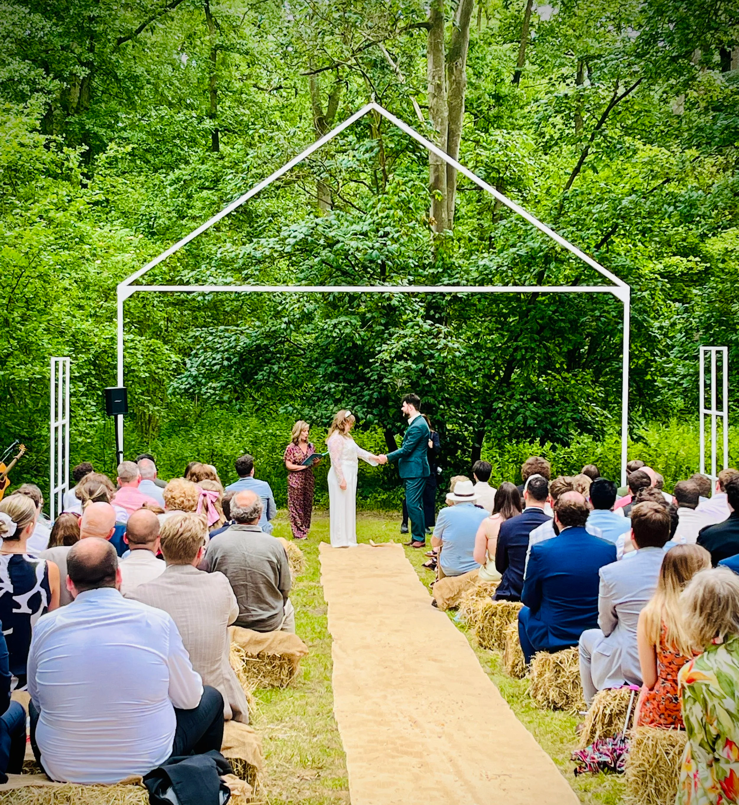 Woodland wedding with a bride and groom getting married in a woodland setting with tall trees and guests seated on straw bales at Stanford Farm