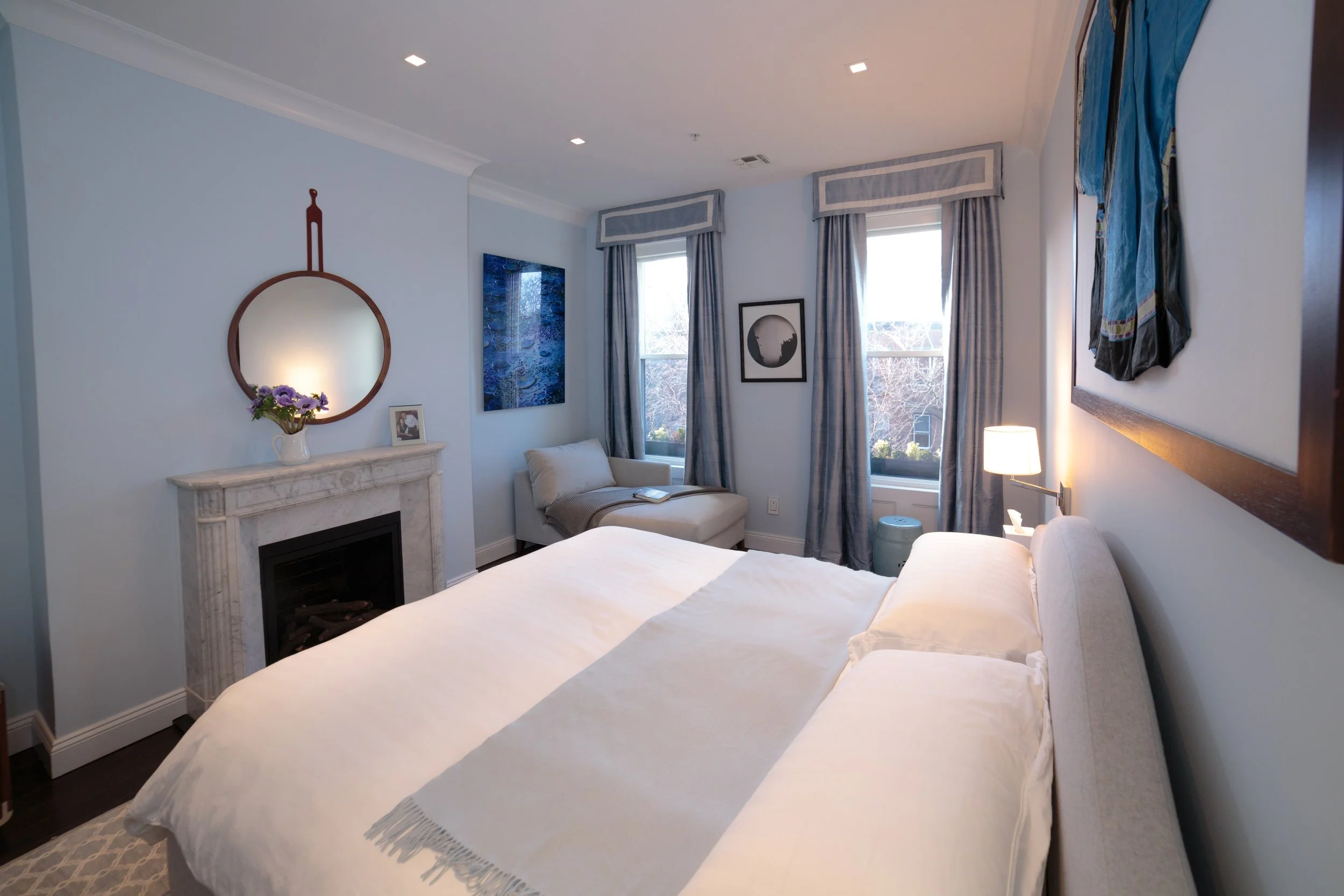A serene light blue master bedroom featuring a white marble fireplace, a round wood-framed hanging mirror, a grey corner chaise lounge, and a bed styled with crisp white and soft blue linens.
