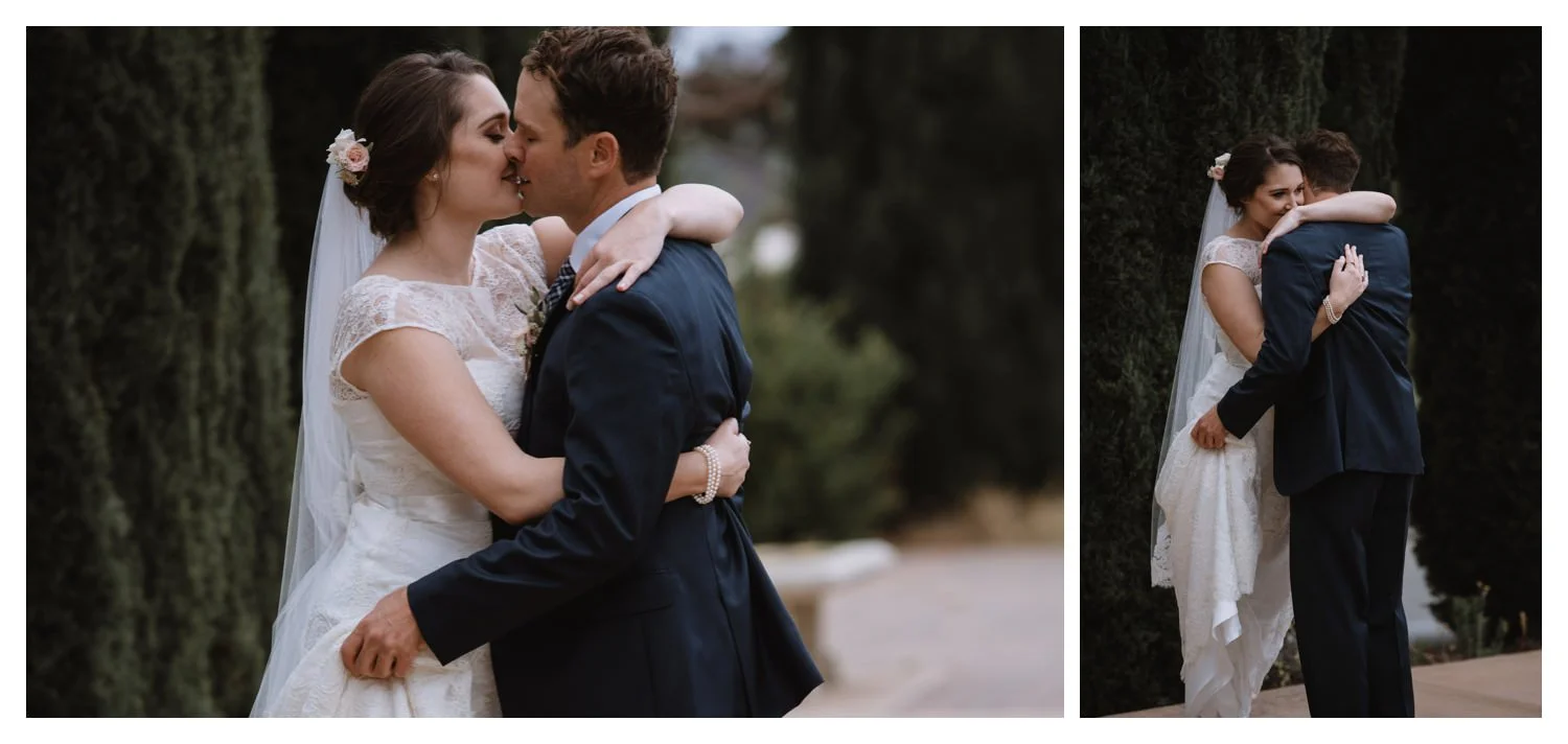 A wedding couple embrace in front of a row of evergreen trees