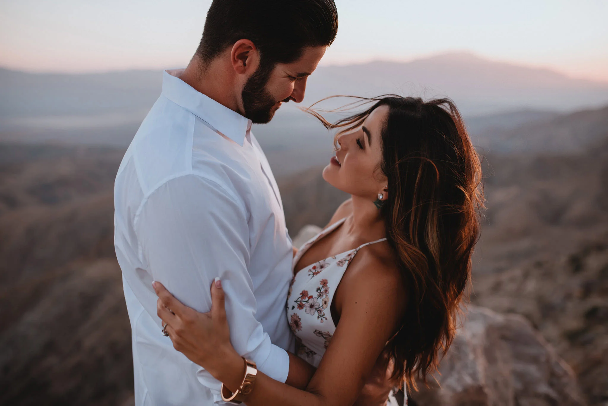 An engaged couple standing on a cliff as the wind blows in Joshua Tree
