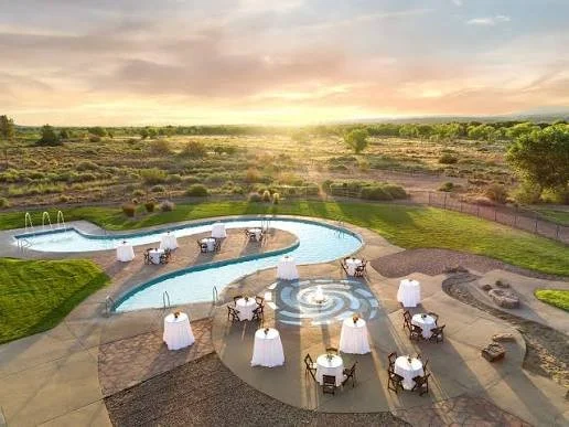 Outdoor swimming pool with lounge chairs and tables with umbrellas on a patio, surrounded by open fields and a sunset sky.