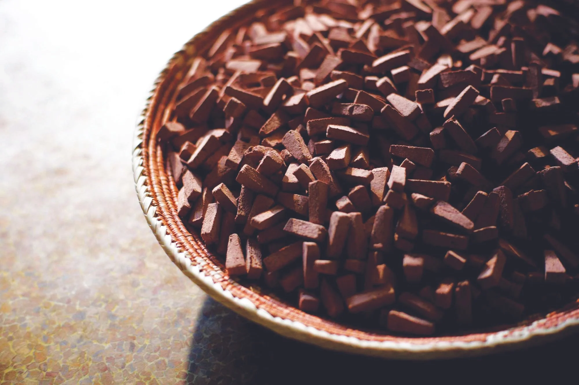 A close-up of a woven basket filled with small blocks of dark chocolate on a textured surface.