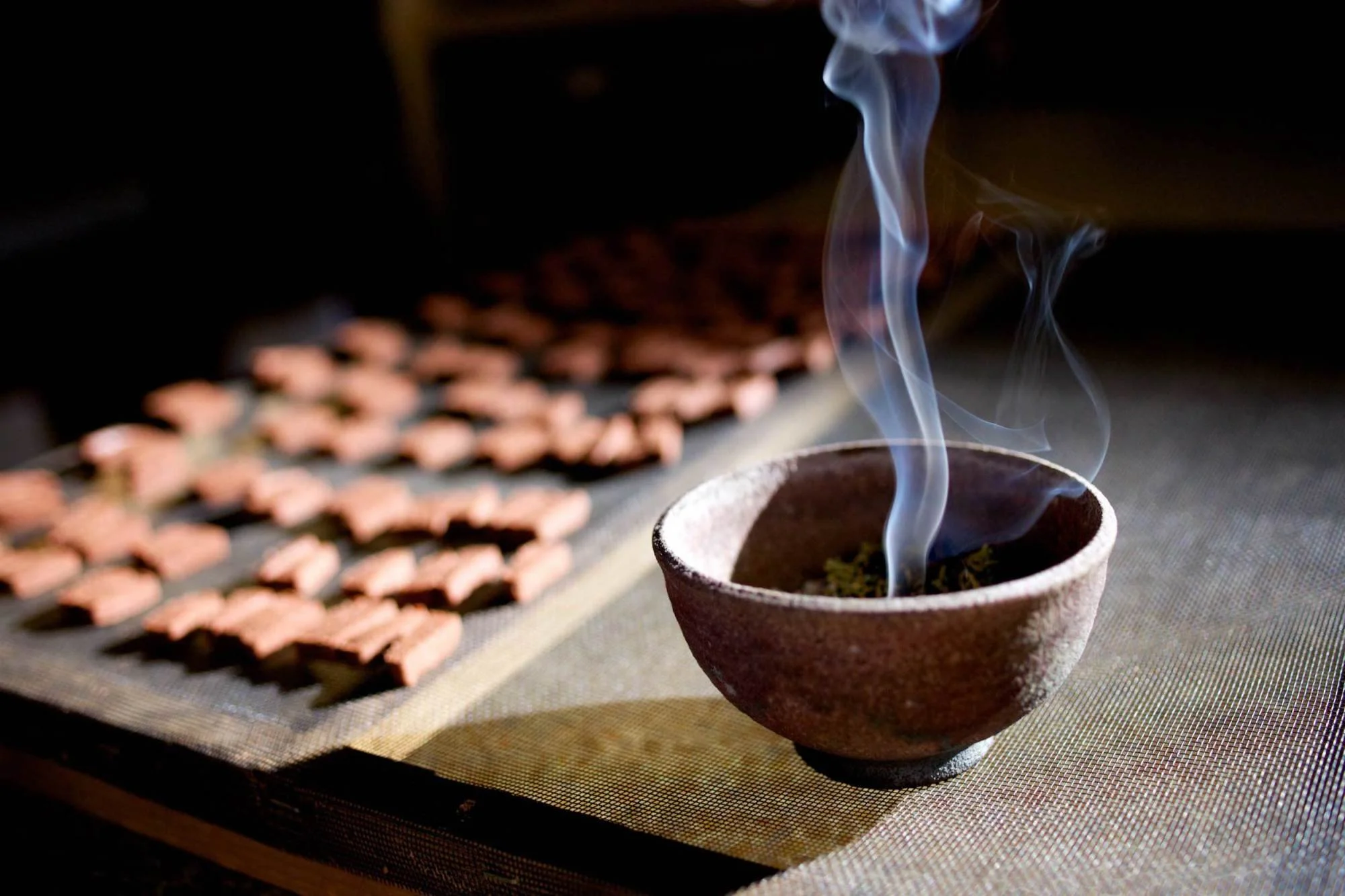 A small ceramic bowl with smoke curling from inside, resting on a textured mat. In the background, there are numerous small, reddish-brown rectangular objects arranged in rows on a cooling rack.