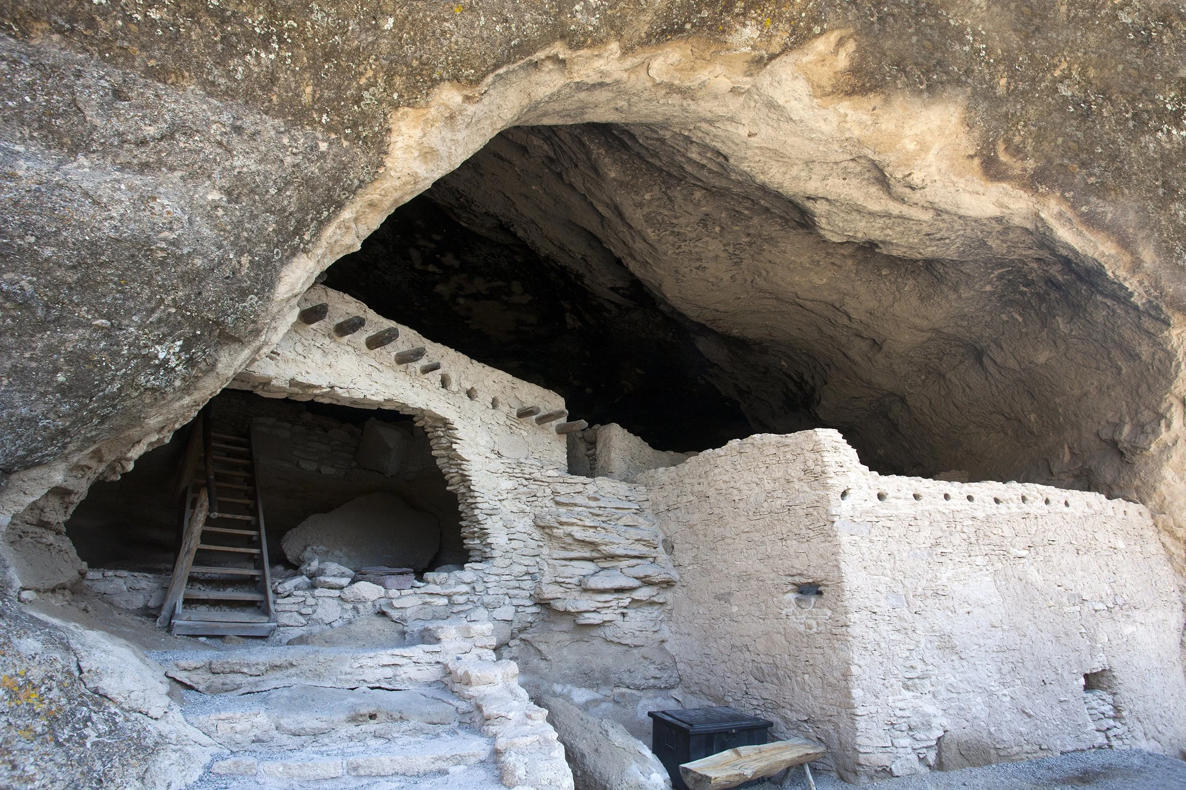 Ancient stone cliff dwelling with a multi-level structure built into a cave, featuring stairs and small windows, with a rocky ceiling overhead.