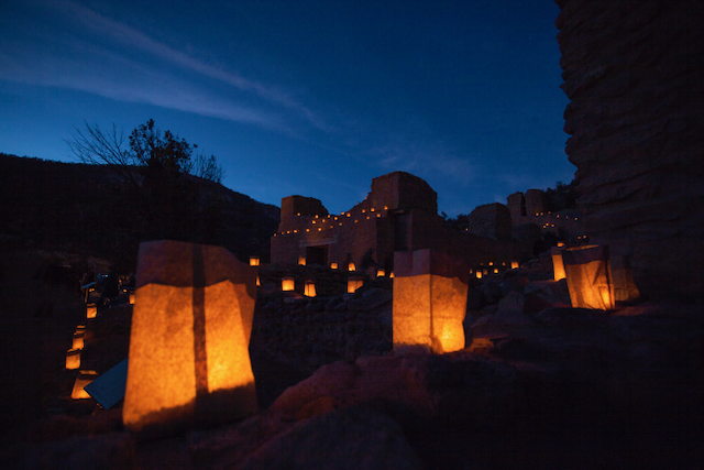 Nighttime view of an ancient ruins site illuminated by lanterns, with stone structures and a deep blue sky.