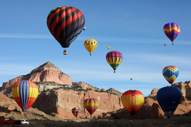 Multiple hot air balloons in various colors floating over a rocky desert landscape with red and beige cliffs under a clear blue sky.