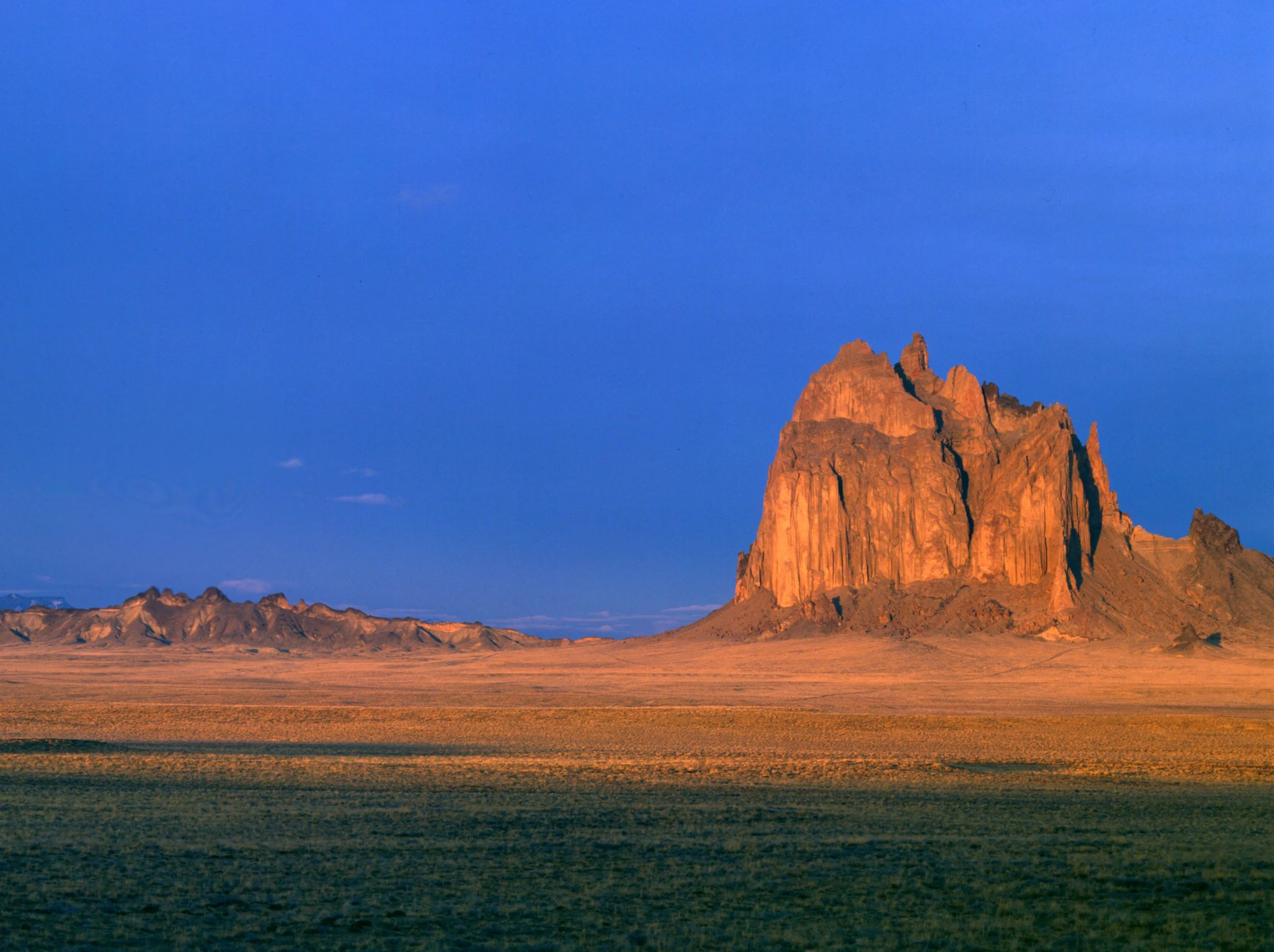 A large desert rock formation under a blue sky with minimal clouds, surrounded by flat, arid land.