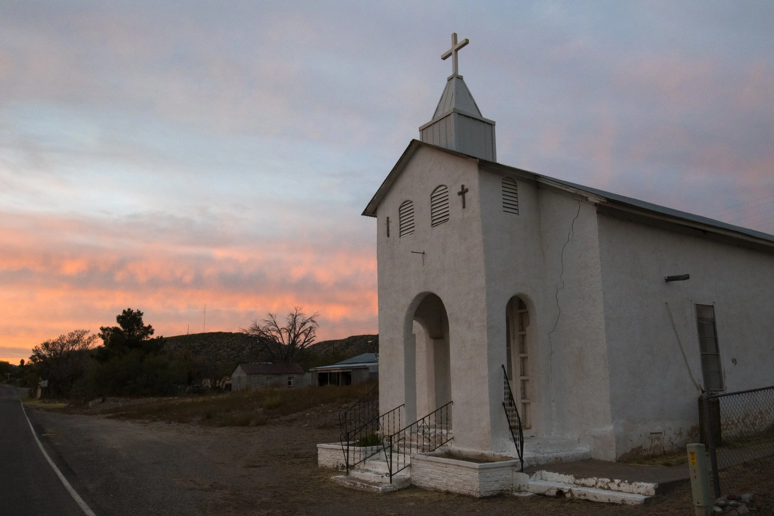 A small white church with a cross on top, situated along a rural road at sunset with pink and blue clouds and hills in the background.