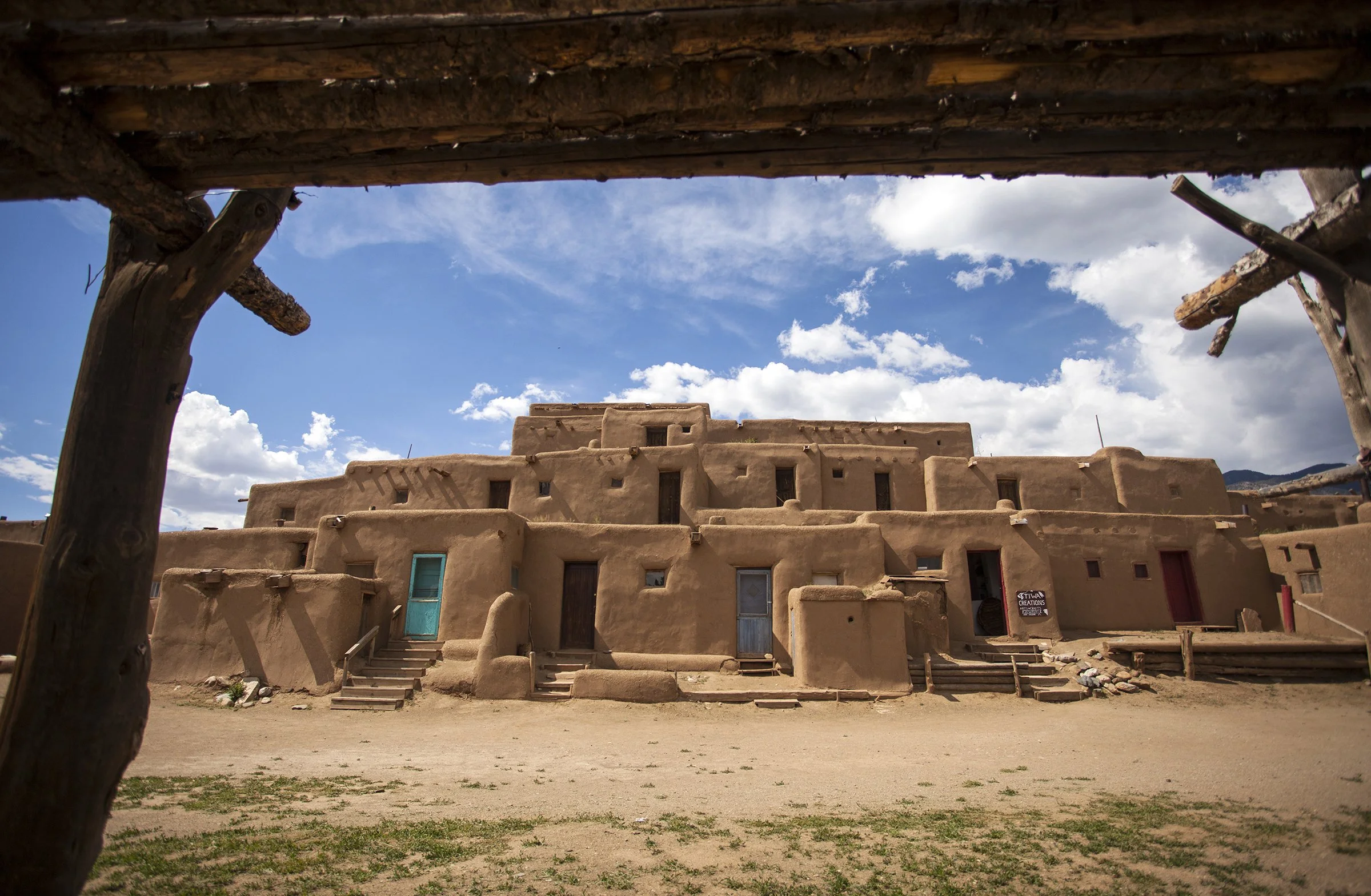 Traditional adobe-style buildings on a sunny day, seen through a wooden frame with a sandy ground and partly cloudy sky.