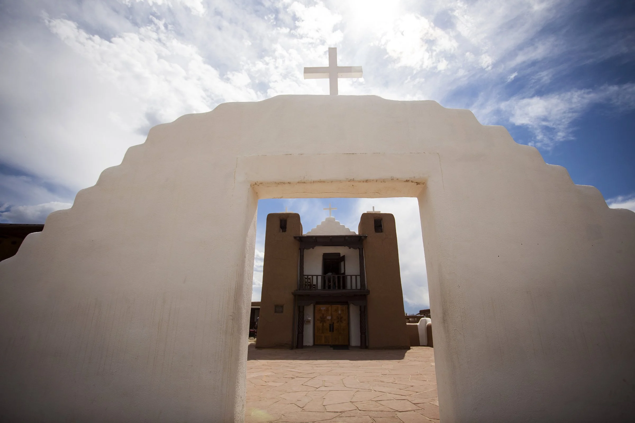 A white adobe church with a cross on top, viewed through an arched entrance, with a second smaller cross visible in the background against a partly cloudy sky.