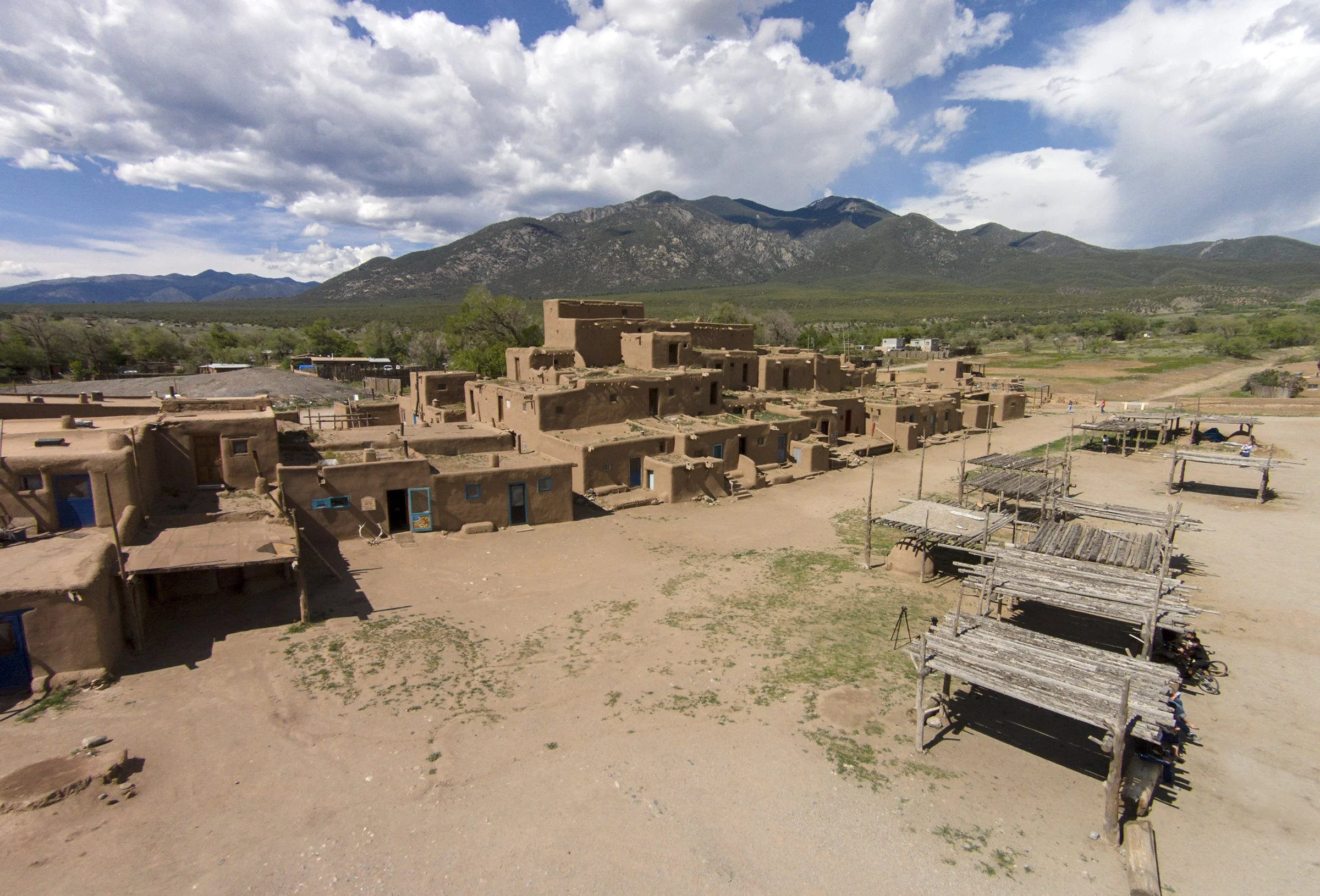 Aerial view of a historic pueblo with adobe-style homes, wooden structures, and mountains in the background under a partly cloudy sky.