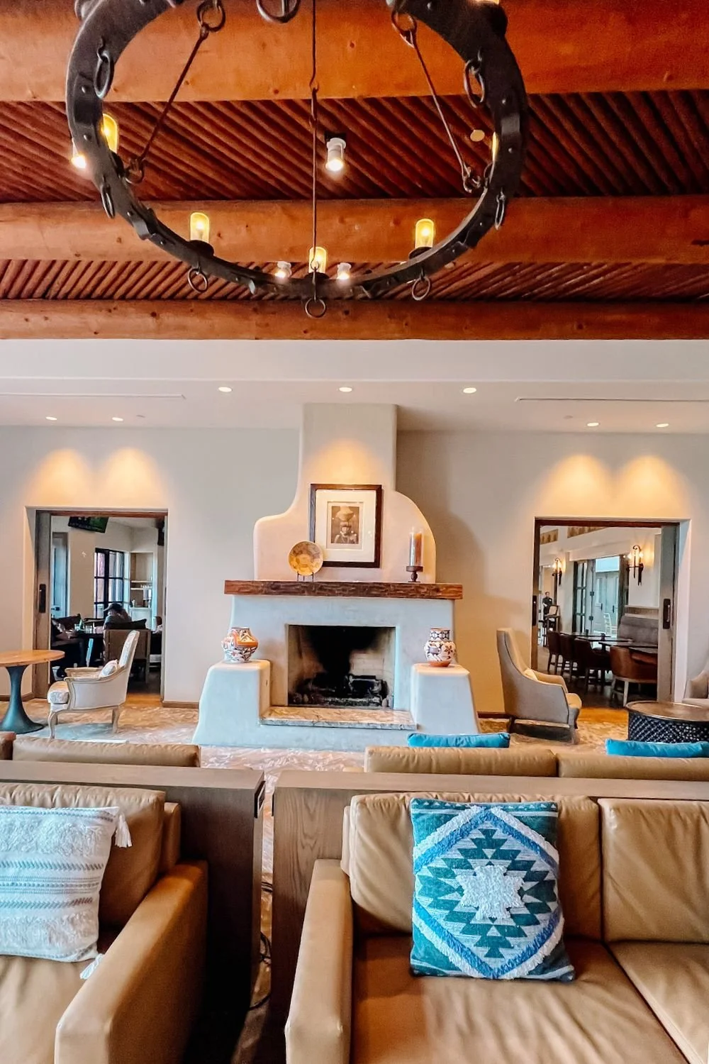 Interior of a cozy living room with a white fireplace, wooden mantel, and decorative vases. There are leather sofas with patterned cushions, and a large horseshoe-shaped chandelier hanging from a wooden ceiling. Two doorways lead to other seating areas, featuring armchairs and tables.