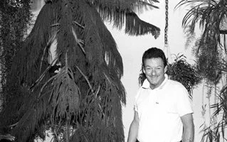 Joshua Peine  smiling outdoors, standing next to a large palm tree, with other plants and trees in the background.