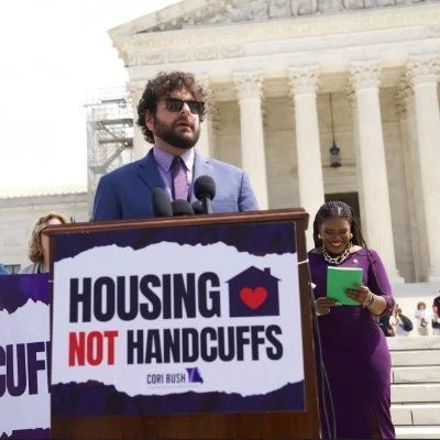 Image is of a person standing behind a podium with microphones speaking to an audience out of view. On the front of the podium is a sign with text reading, "Housing Not Handcuffs." The speaker is wearing a suit and tie and sunglasses.
