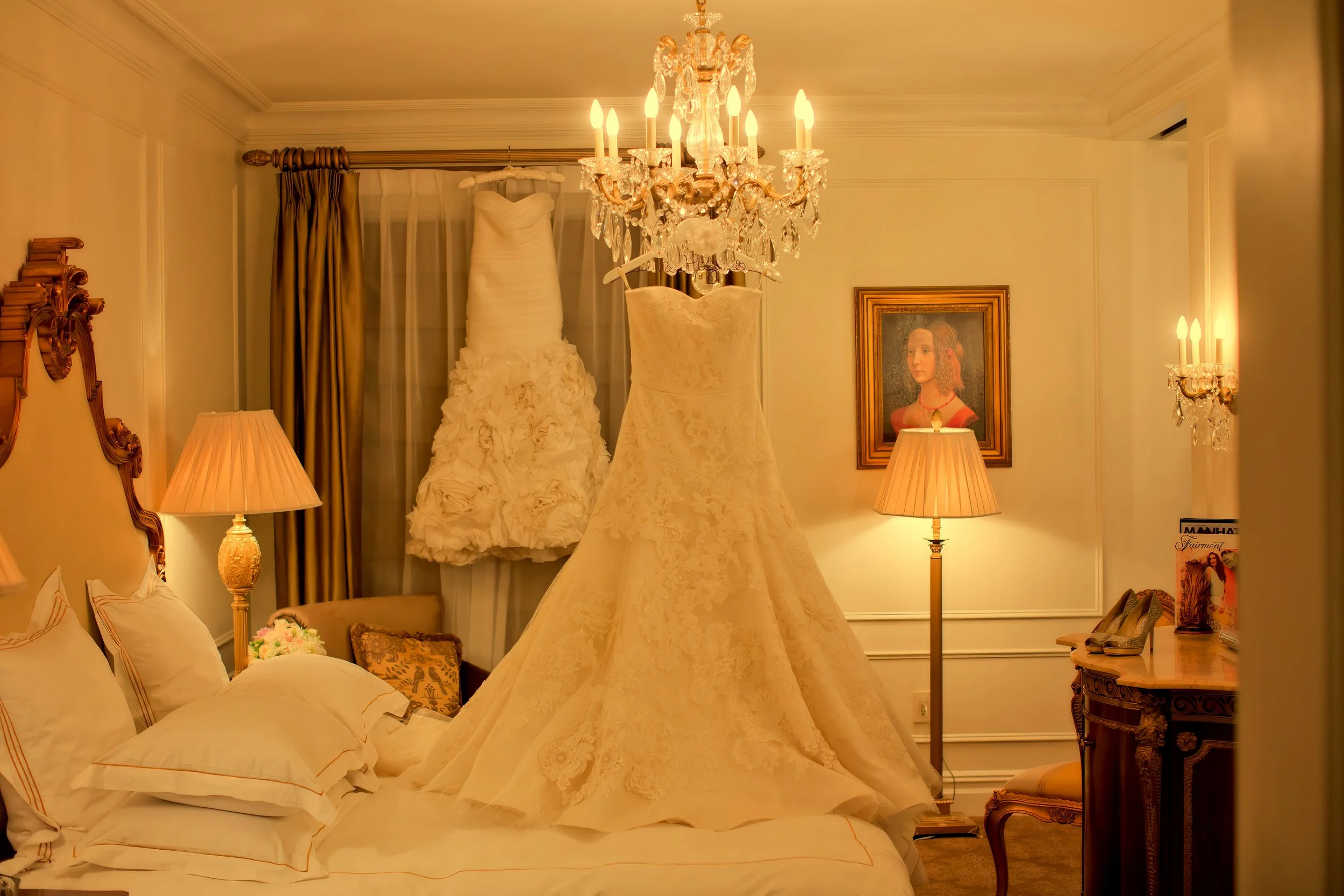Two elegant wedding gowns displayed beneath a crystal chandelier inside a luxury suite at The Plaza Hotel in New York.