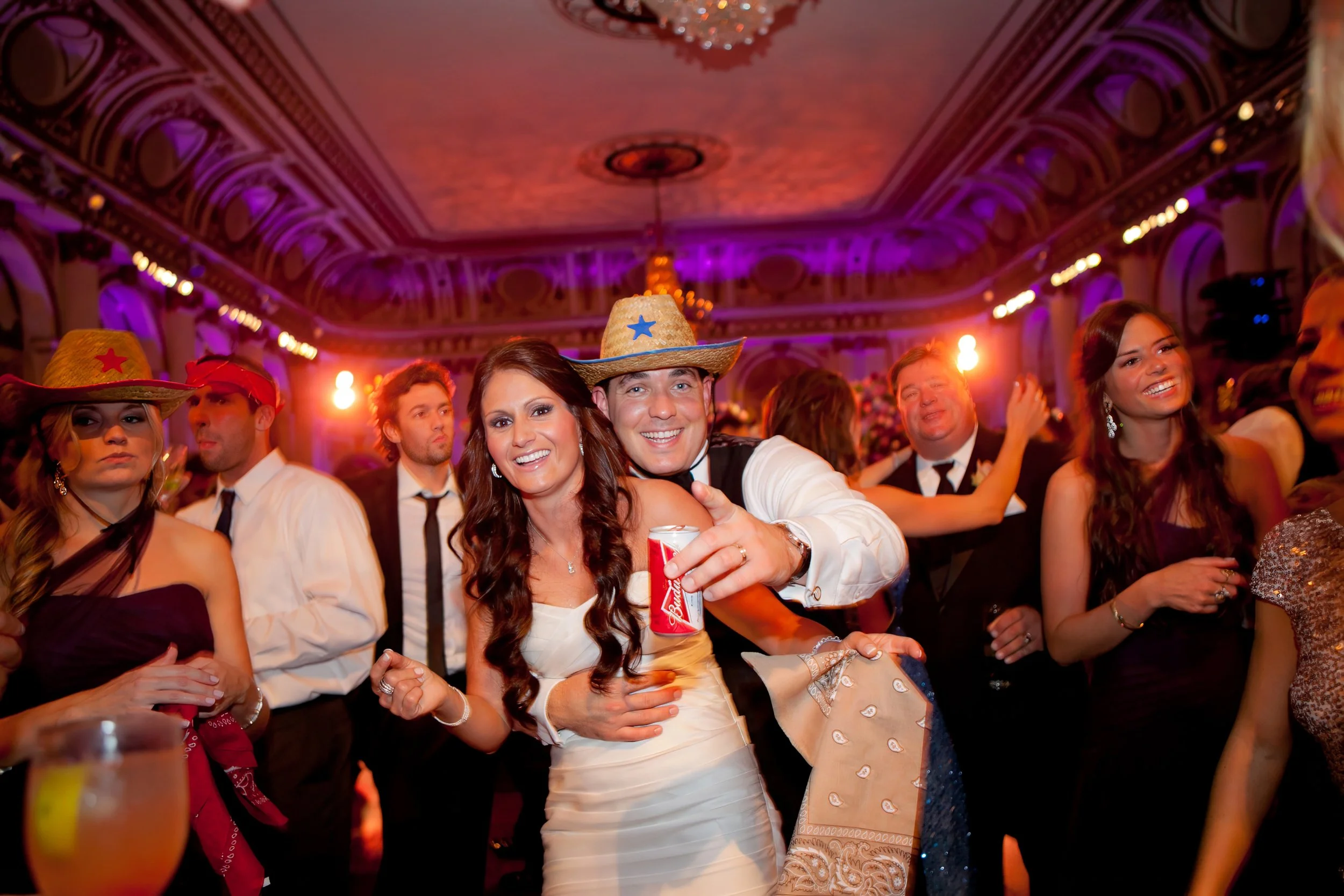 Bride and groom smiling with wedding guests wearing playful cowboy hats during a lively Plaza Hotel reception.