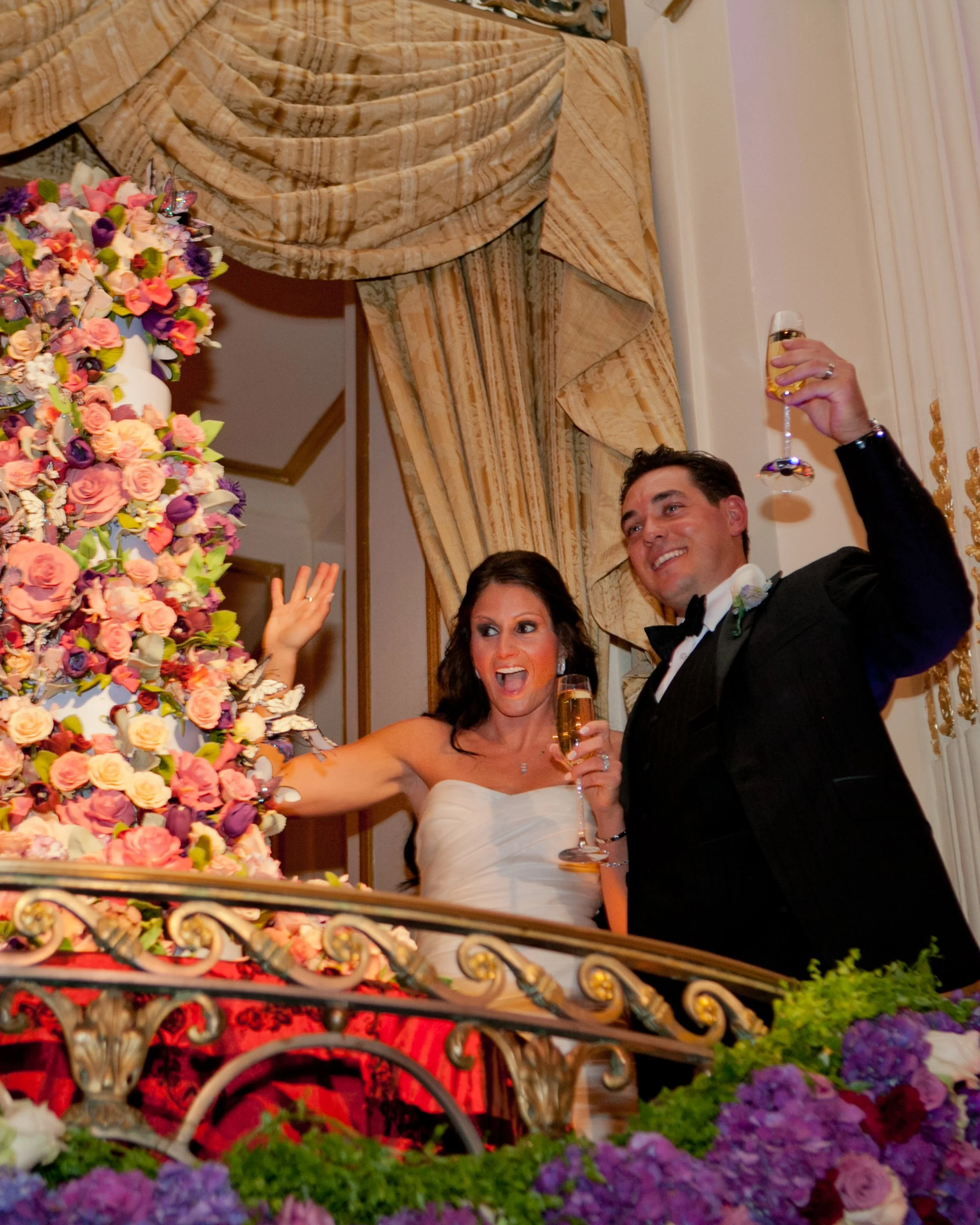 Bride and groom raise champagne glasses beside an elaborate floral wedding cake during their reception at The Plaza Hotel.