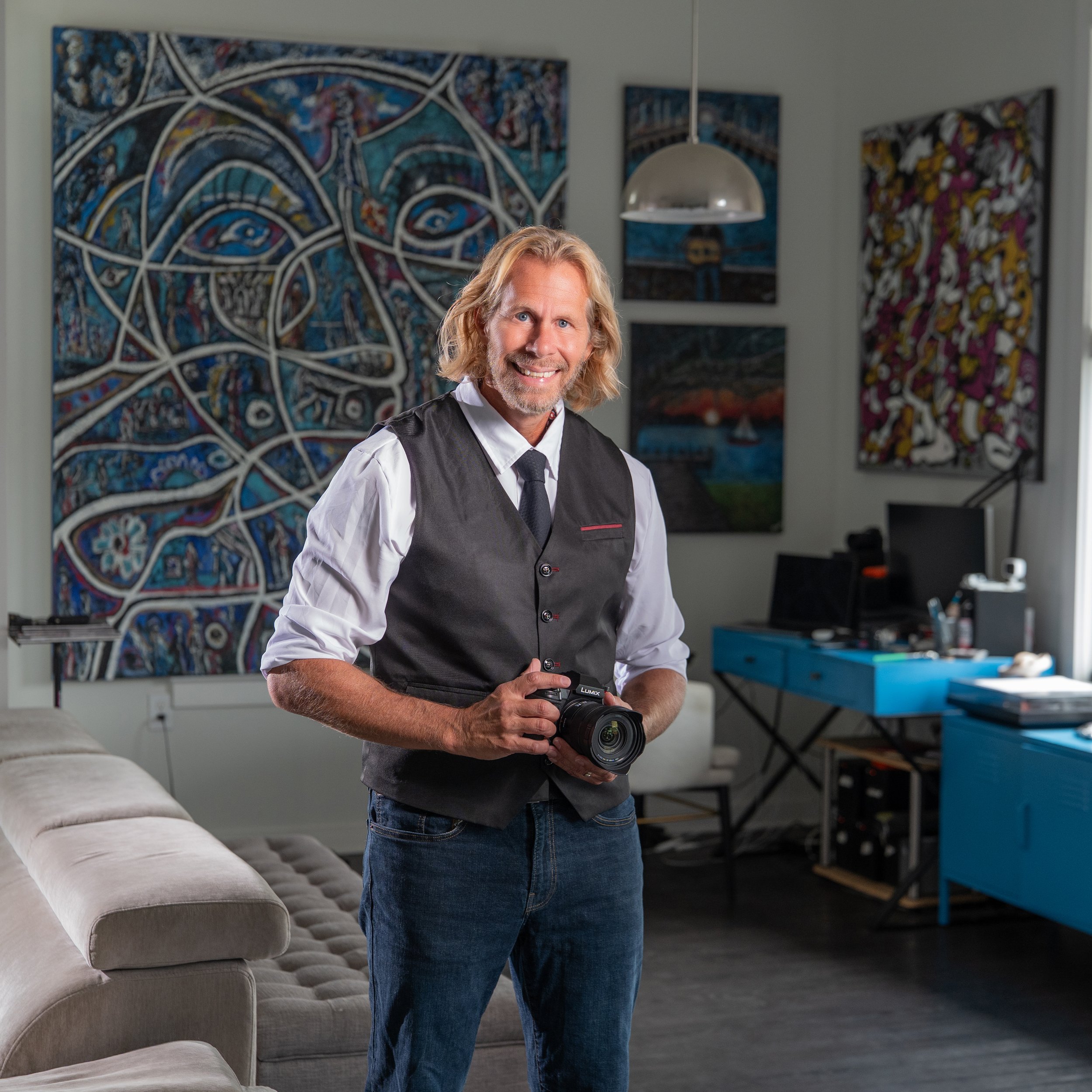 Charles Maring smiling and holding a camera in his Savannah art studio, surrounded by original abstract paintings.