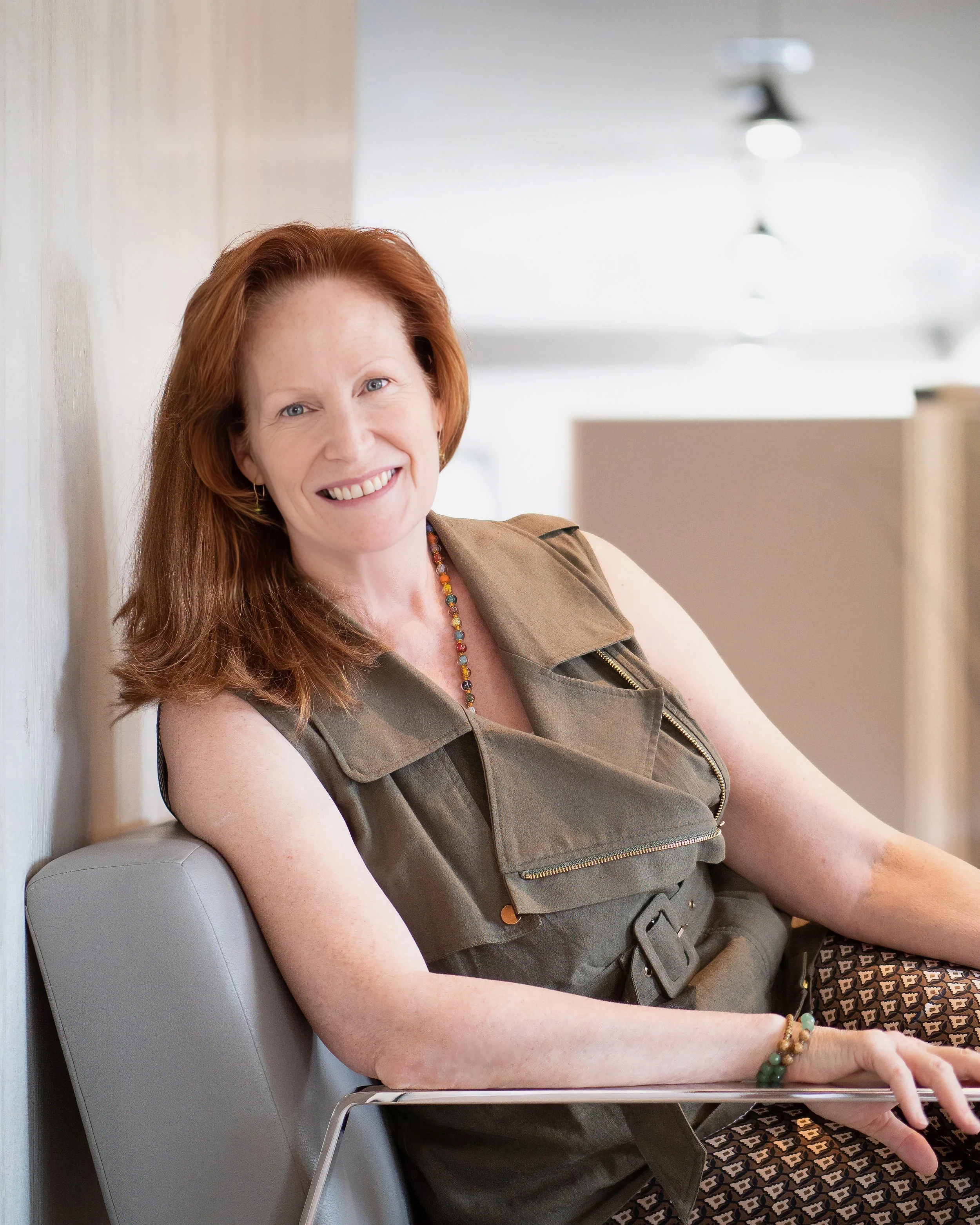 Red-haired professional woman smiling while seated comfortably in a modern chair, wearing an olive green sleeveless top.