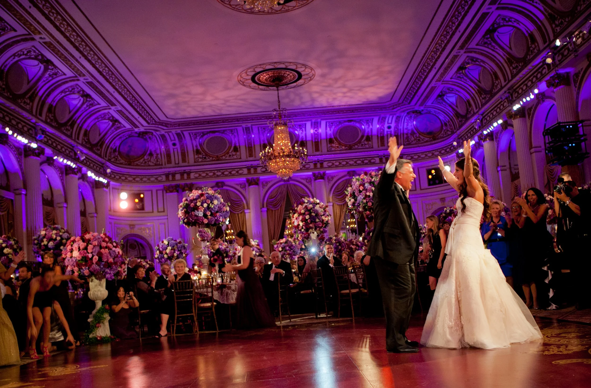 Bride and her father raise their hands toward each other during an energetic dance beneath purple ballroom lighting at The Plaza Hotel.