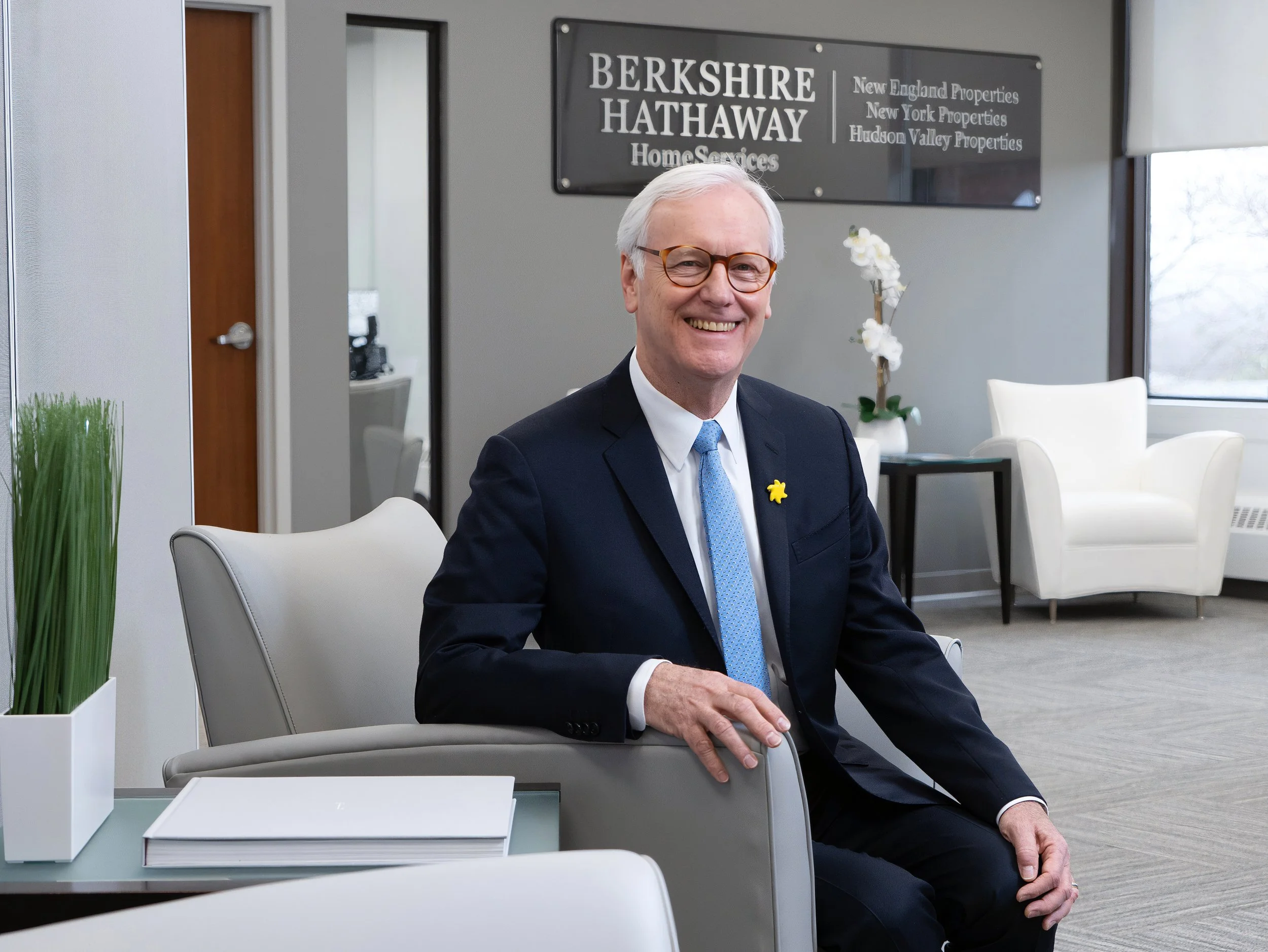 Smiling Berkshire Hathaway HomeServices executive in a dark suit and blue tie, seated in a modern office with company signage visible behind him.