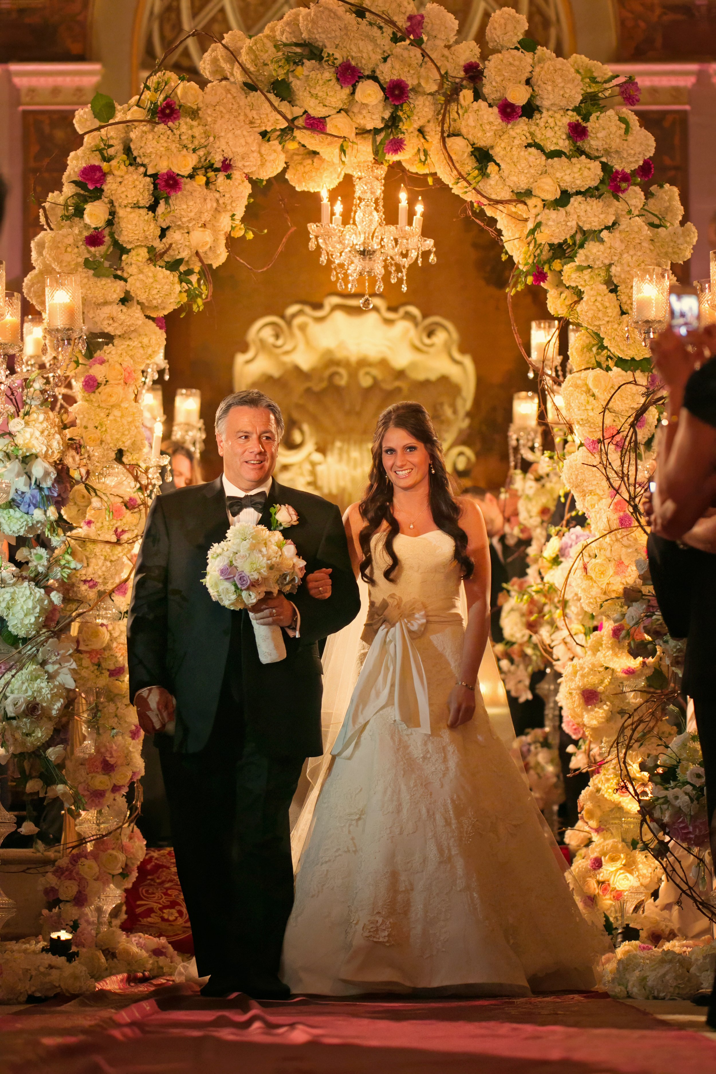 Bride walking arm in arm with an escort beneath an elaborate floral arch during a candlelit wedding ceremony at The Plaza Hotel.