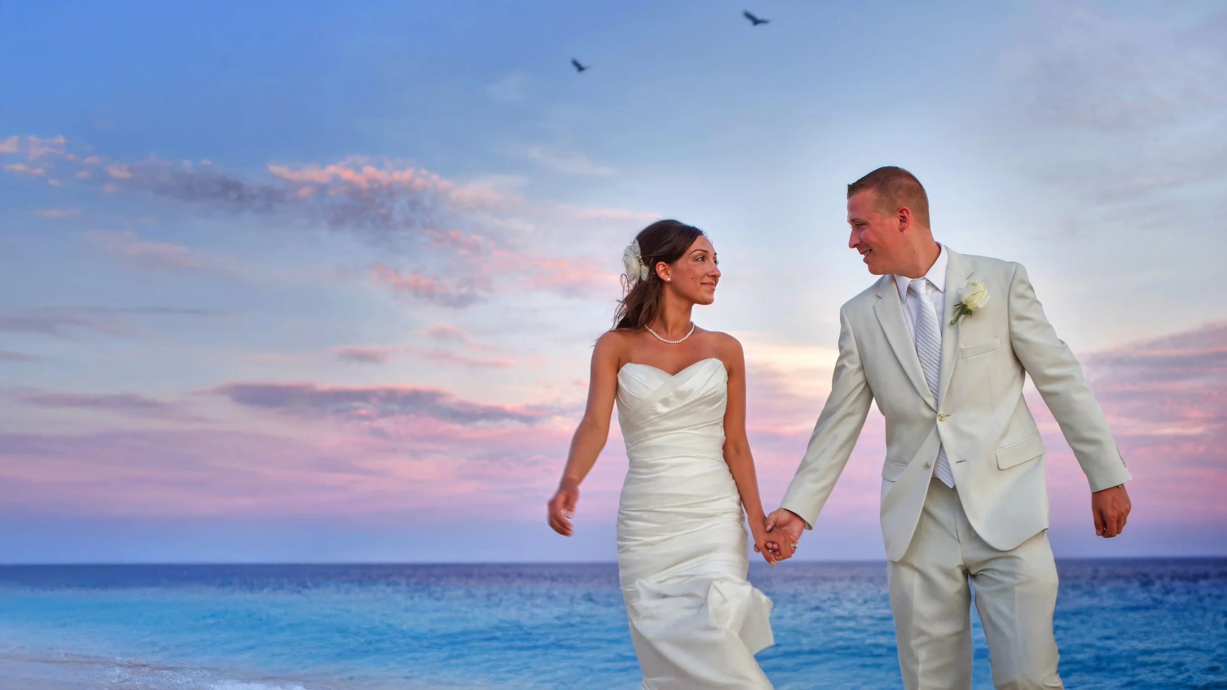Bride and groom walk together along the shoreline in Cabo San Lucas, Mexico, beneath a lavender evening sky with birds flying overhead.