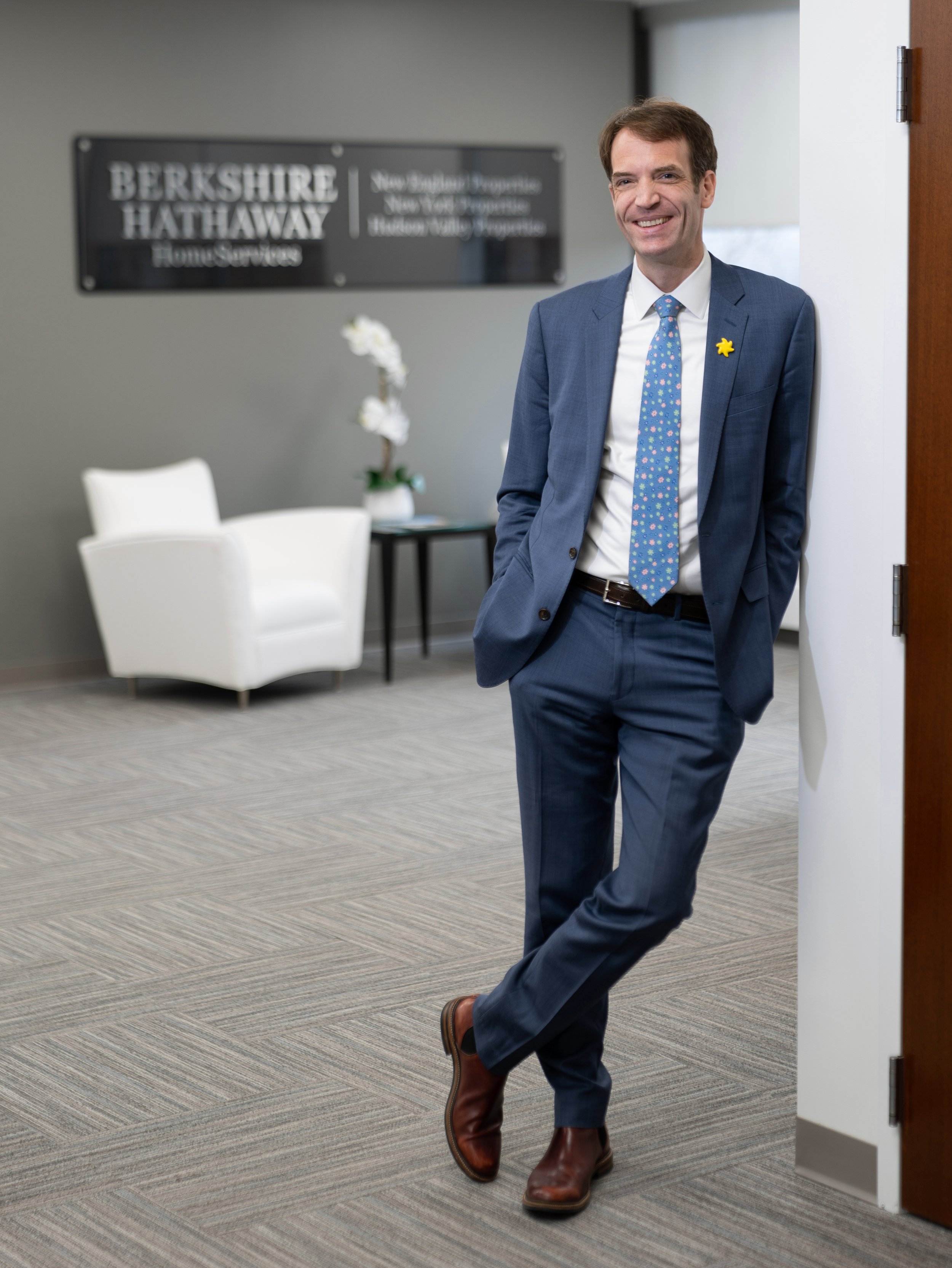 Berkshire Hathaway HomeServices executive wearing a blue suit, leaning casually against an office doorway with the company sign in the background.