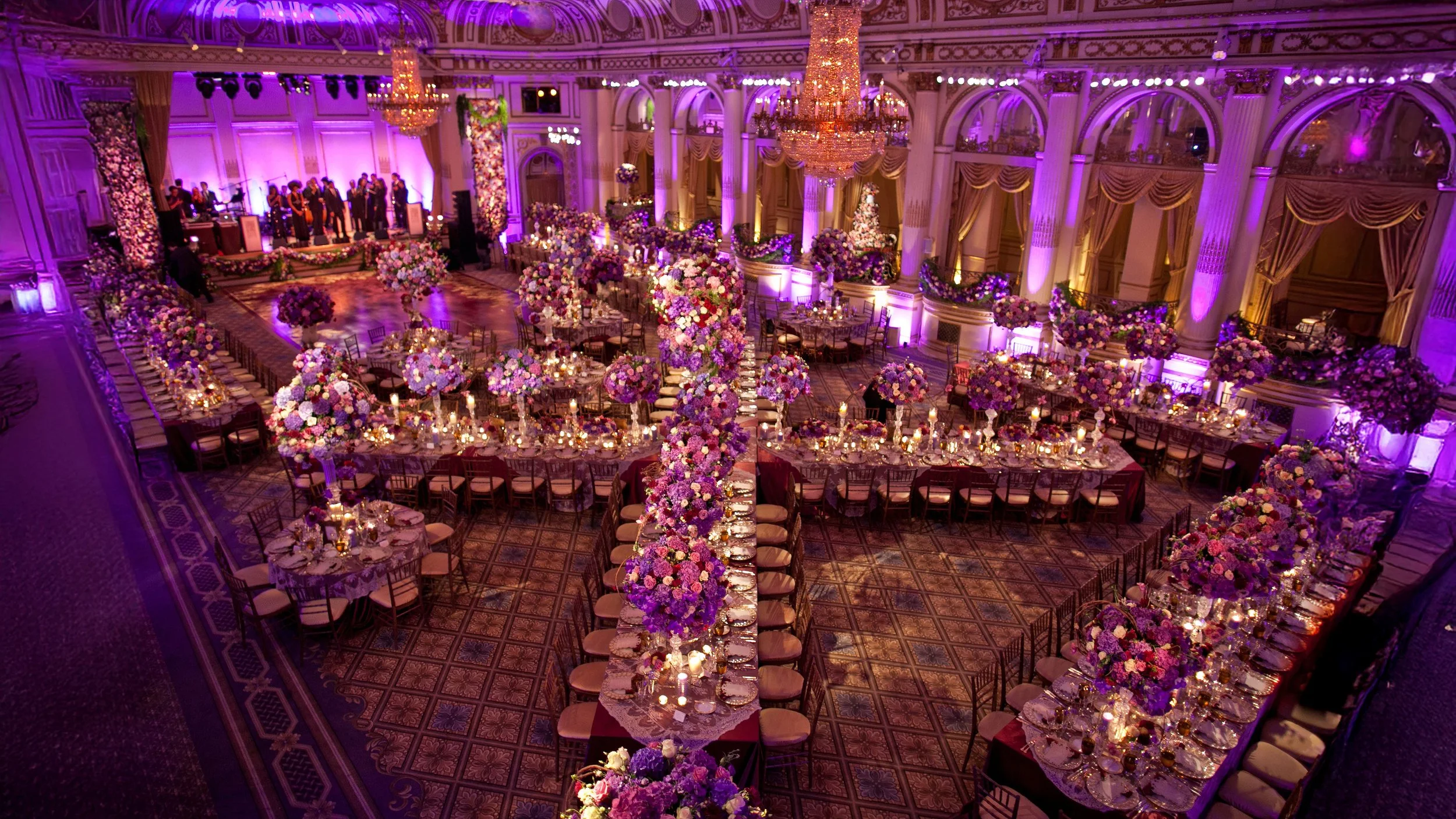 Elevated view of The Plaza Hotel ballroom decorated with purple floral arrangements, candlelit tables, and dramatic lighting.