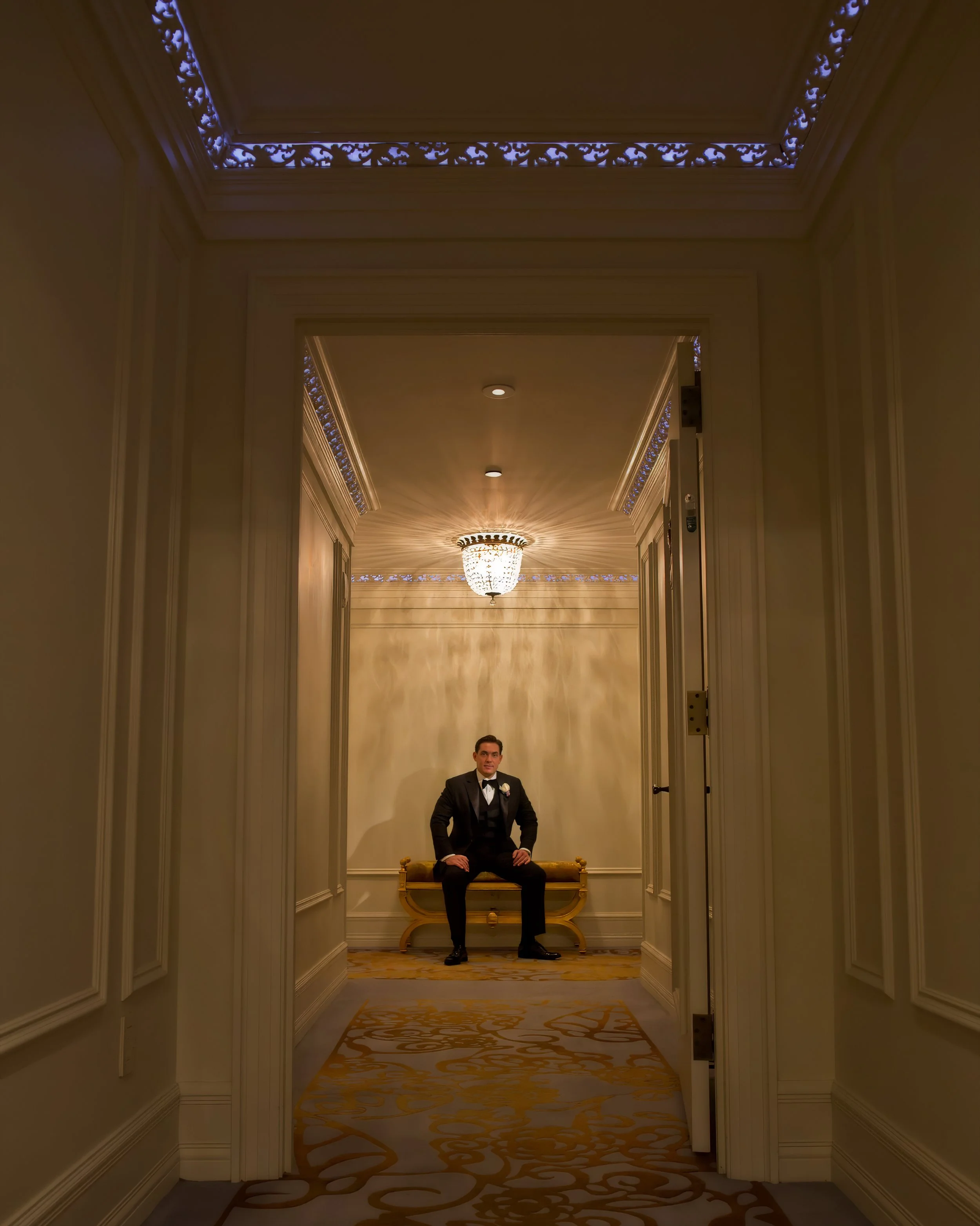 Groom in a black tuxedo seated on a gold bench, framed by a series of elegant doorways inside The Plaza Hotel.