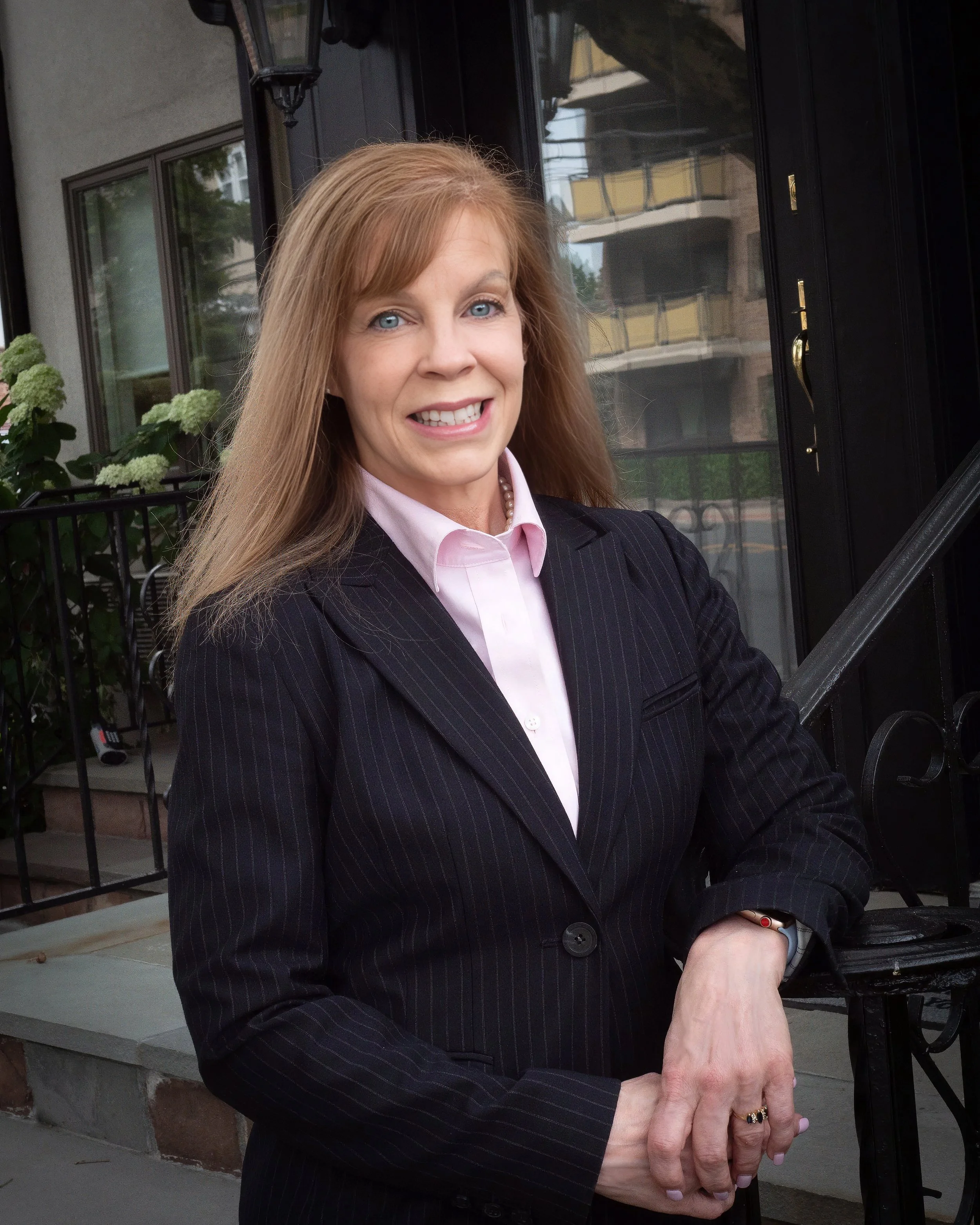 Professional woman wearing a dark pinstripe suit and pink blouse, photographed beside a black railing outside a building entrance.