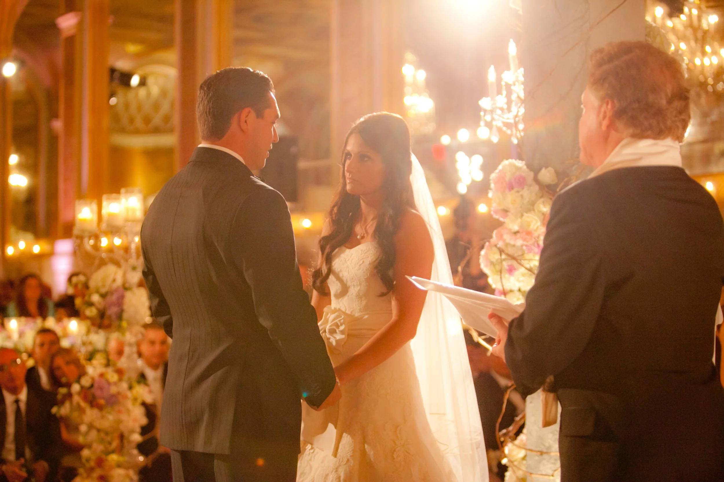 Bride and groom exchange vows during an intimate ceremony surrounded by warm light, flowers, and wedding guests at The Plaza Hotel.