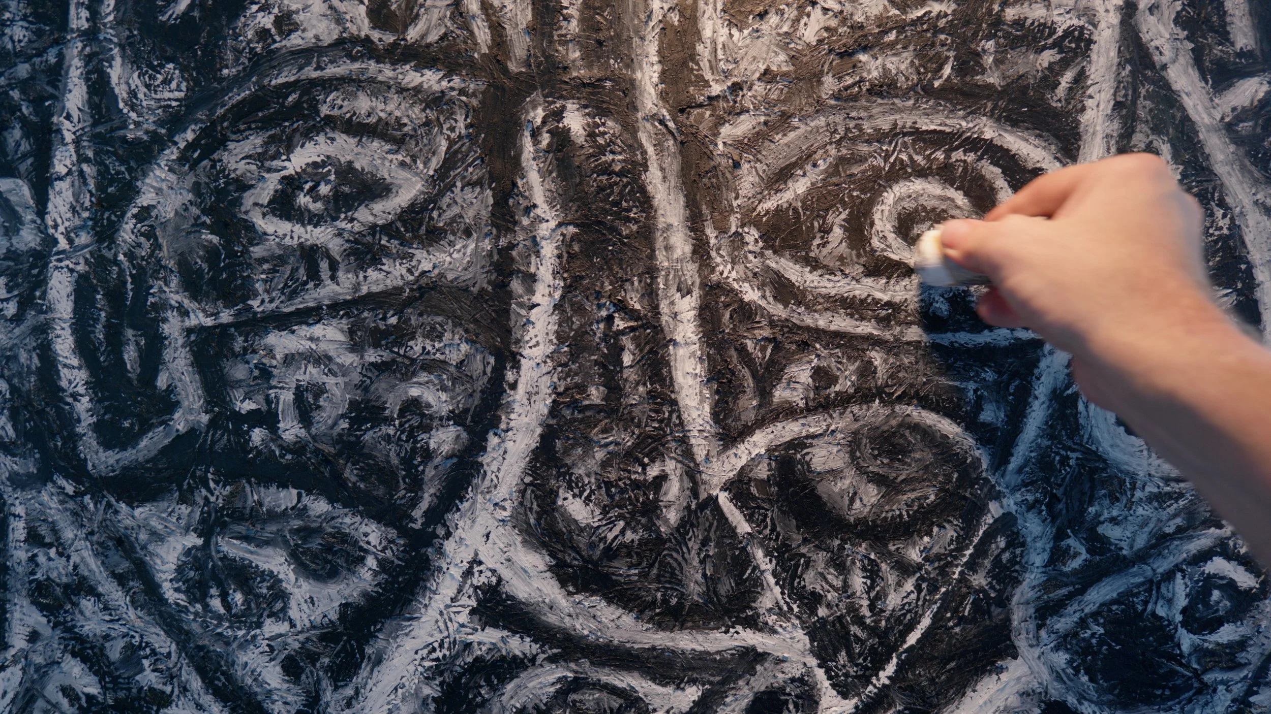 Charles Maring’s hand applying paint to a deeply textured artwork as faces and abstract forms emerge from the surface.