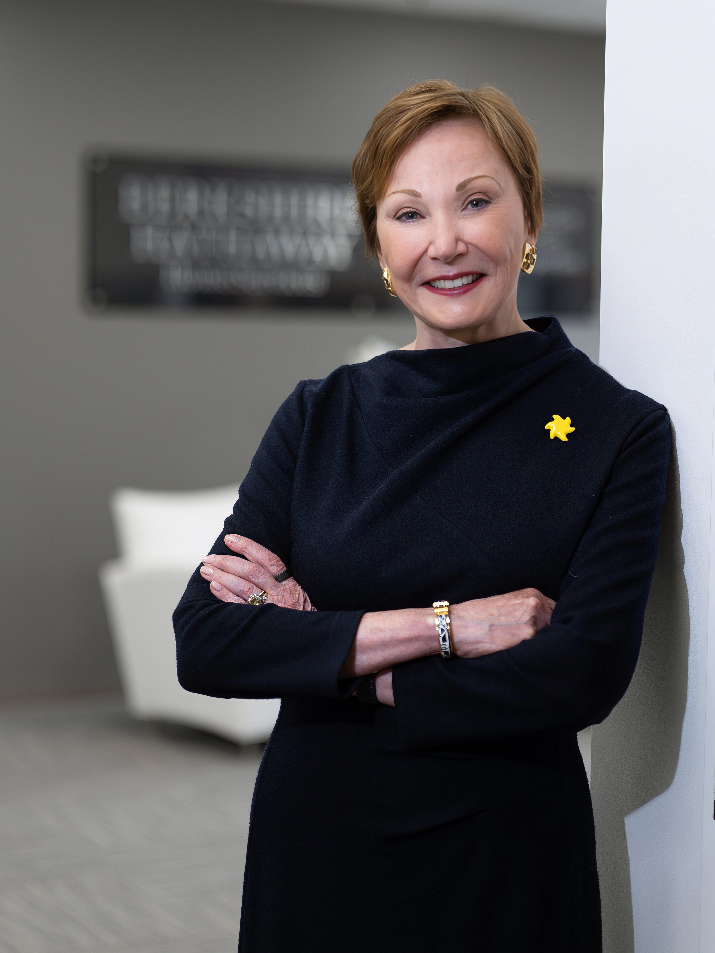 Female Berkshire Hathaway HomeServices executive in a navy outfit, photographed with folded arms against a softly blurred corporate office background.