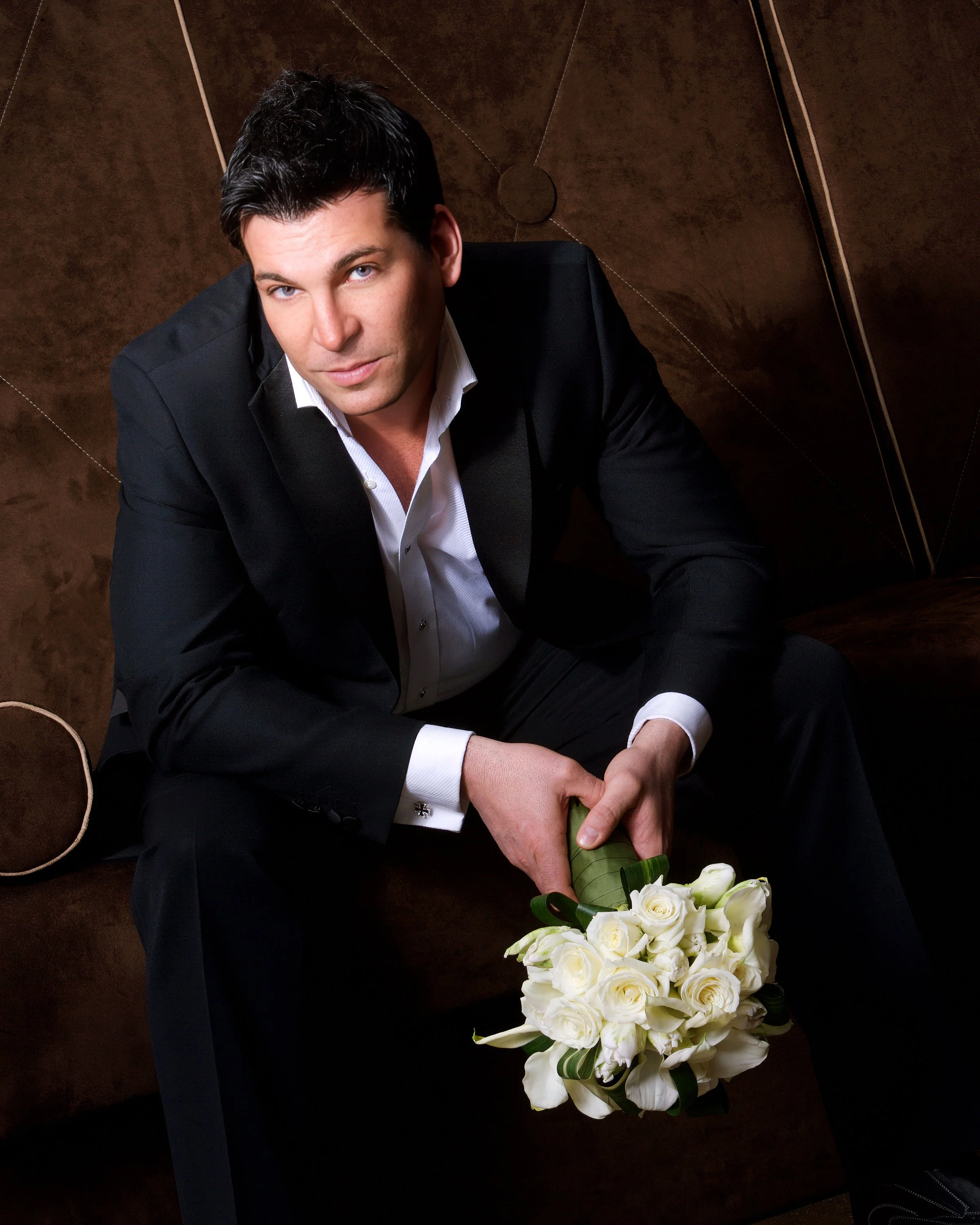 Formal editorial portrait of David Tutera seated in a black suit holding a bouquet of white roses, photographed by Charles Maring.