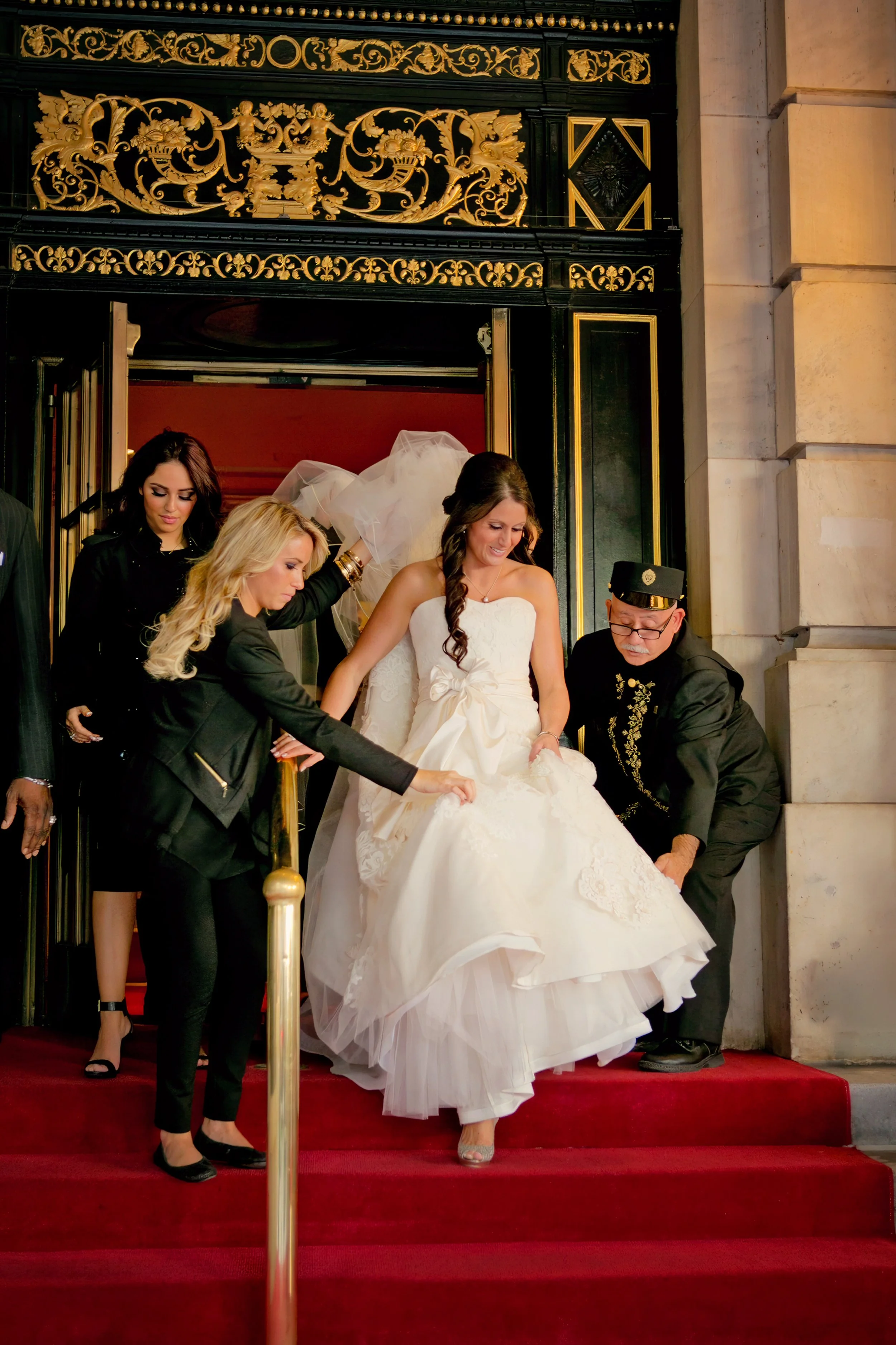 Bride in an elegant wedding gown being assisted down the red-carpeted steps outside The Plaza Hotel by members of the wedding team.