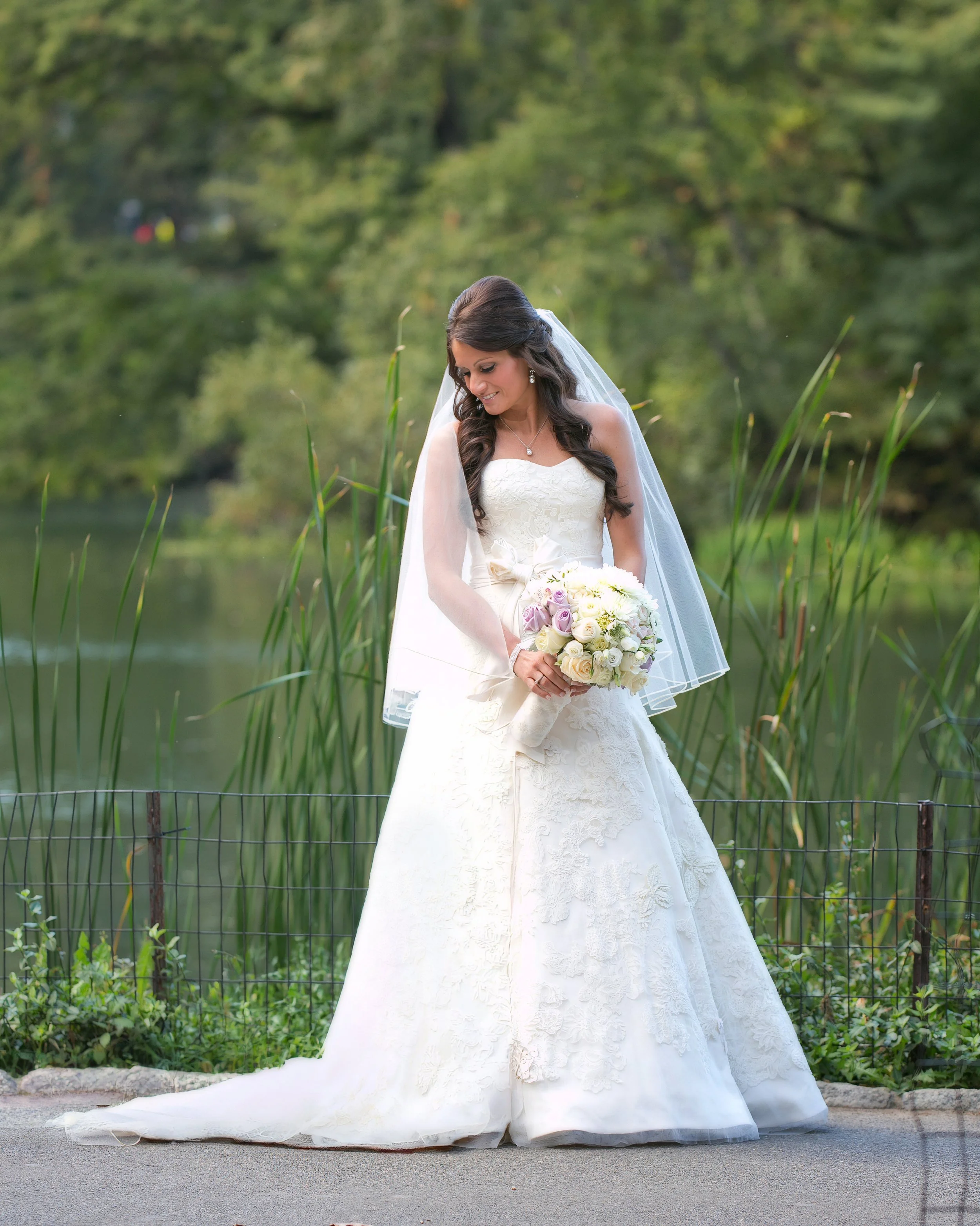 Bride in a white wedding gown and veil holding a pastel bouquet beside a peaceful pond surrounded by greenery.