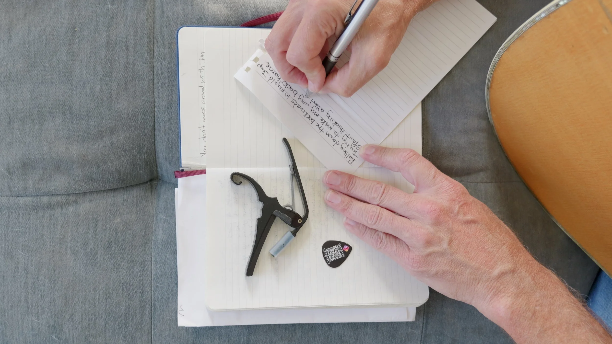 Charles Maring writes lyrics for The Golden Road in a songbook beside an acoustic guitar, capo, and guitar pick.